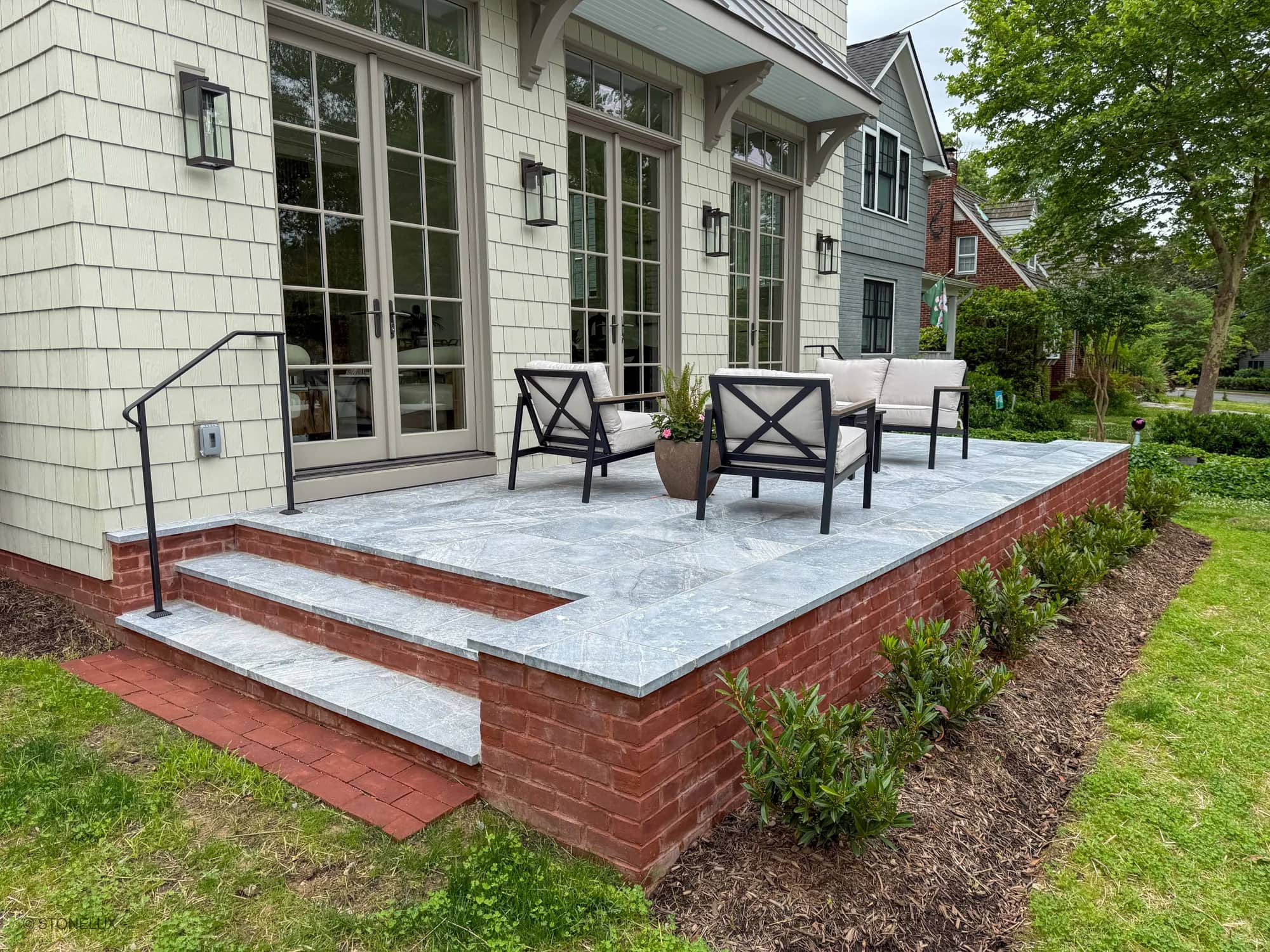 Outdoor patio featuring Aegean Grey Marble Sand Blasted 16x24 3cm Paver by STONELUX. The elegant marble pavers are installed on a raised brick platform, complementing the traditional home architecture with modern outdoor seating and lush greenery.