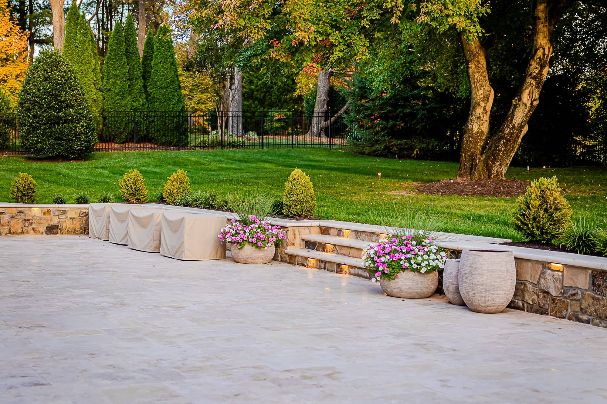 Spacious stone patio, part of the Ivory Twilight: A Travertine Patio Project, features covered seating, colorful planters, softly lit steps, and a landscaped yard with trees and shrubs in the background.