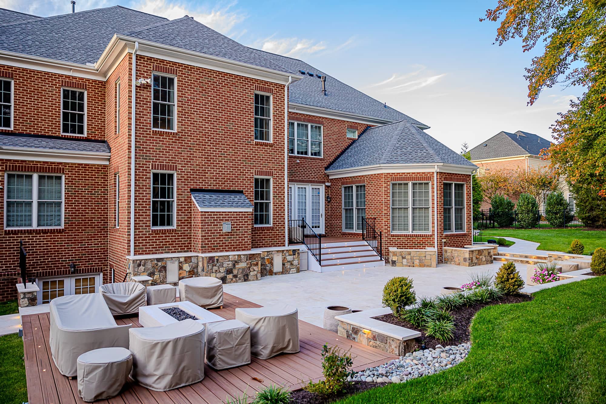 A large red brick house with many windows, a gray roof, and a landscaped backyard featuring an Ivory Twilight: A Travertine Patio Project, outdoor seating with covered furniture, a fire pit, and stone steps leading up to the house.