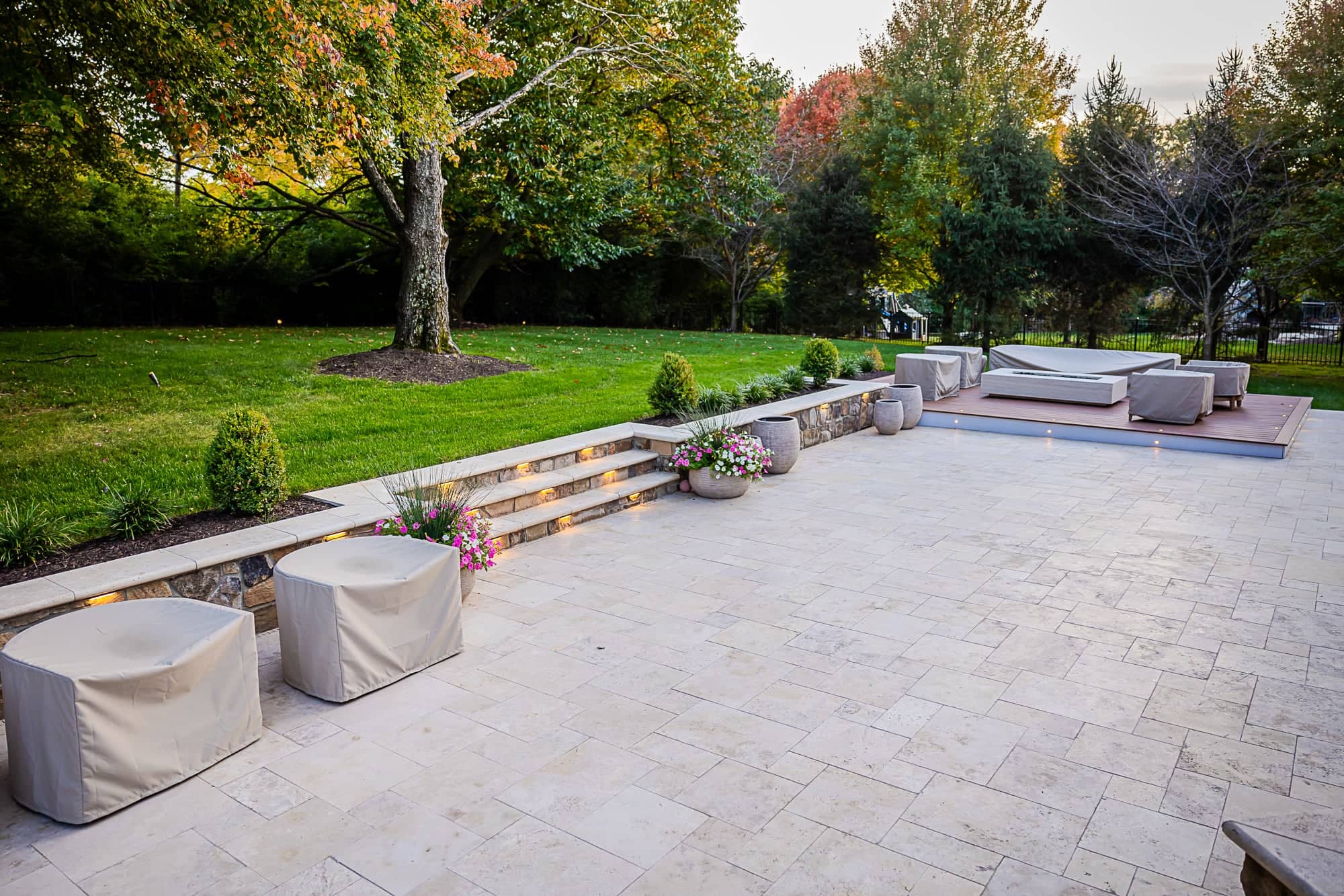 A spacious backyard patio with light stone tiles, part of the Ivory Twilight: A Travertine Patio Project, features covered furniture, potted flowers, and steps with built-in lights, all surrounded by green grass, shrubs, and mature trees in early autumn.