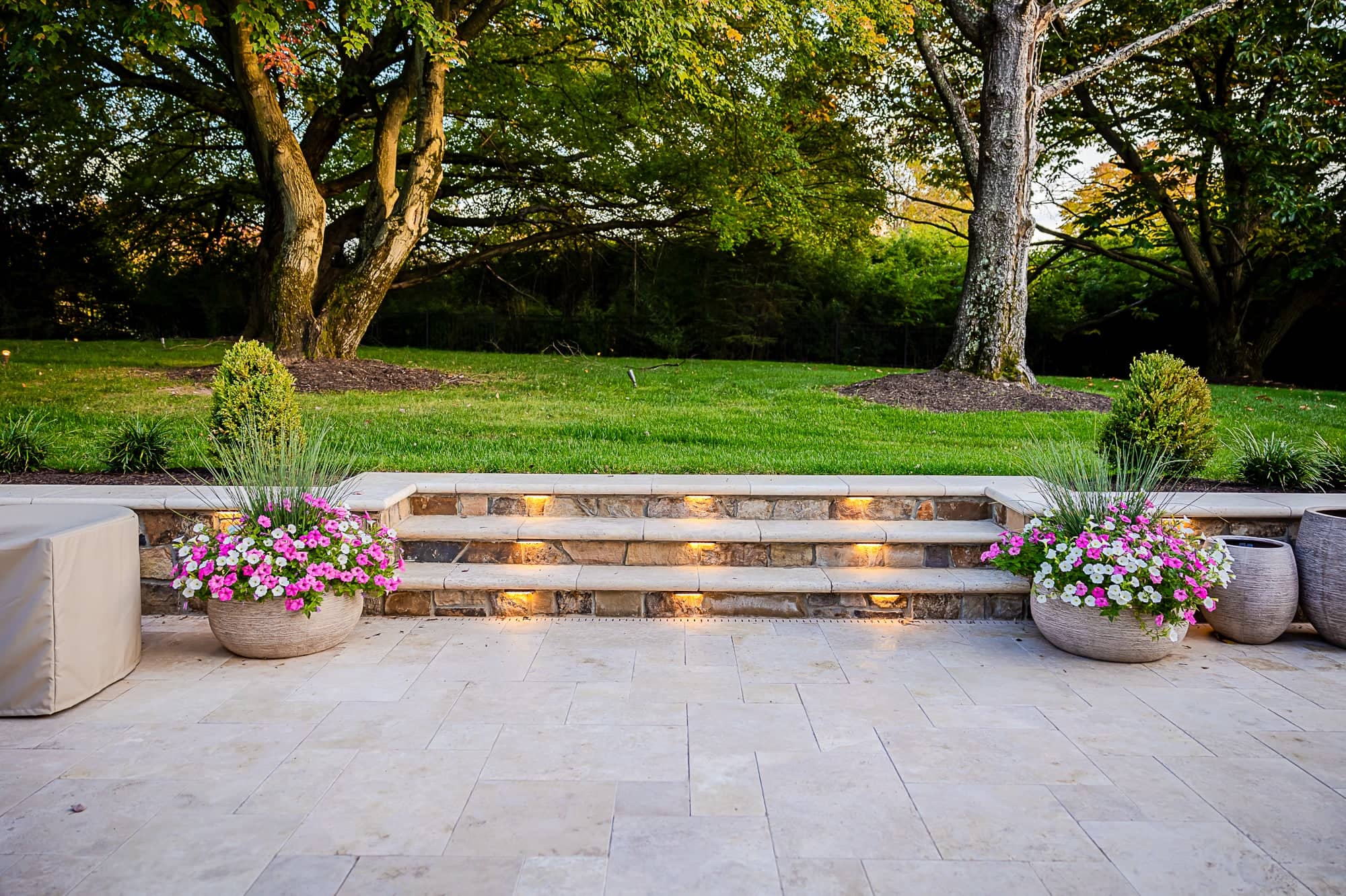Ivory Twilight: A Travertine Patio Project features stone steps with built-in lights leading to a green lawn bordered by trees. Large planters with colorful flowers and tall grass flank the steps, creating a serene outdoor setting.