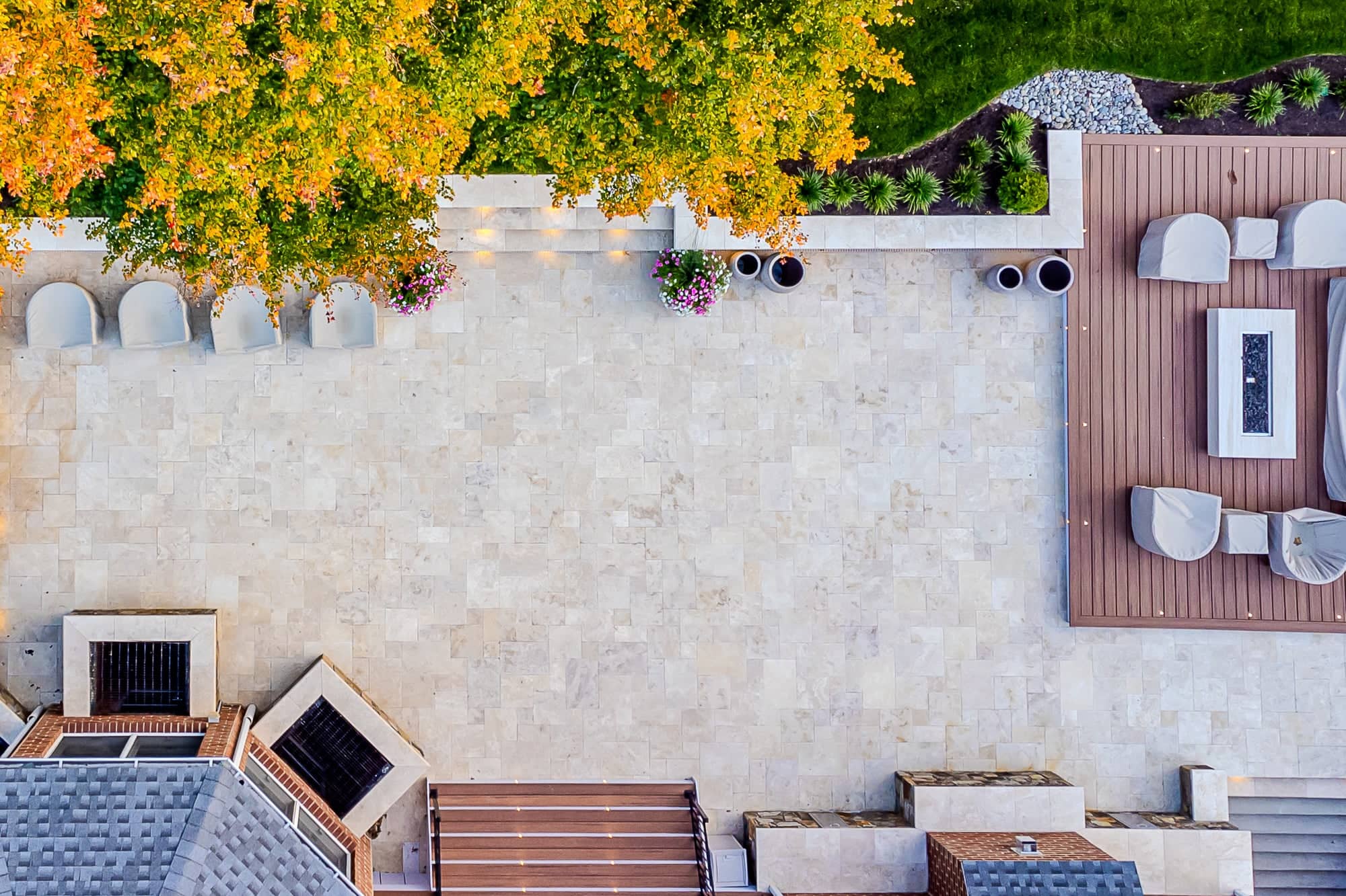 Aerial view of Ivory Twilight: A Travertine Patio Project featuring a stone patio with covered outdoor furniture, fire pit, potted flowers, and lush greenery. A large tree with orange-green leaves casts dappled shade across the area.