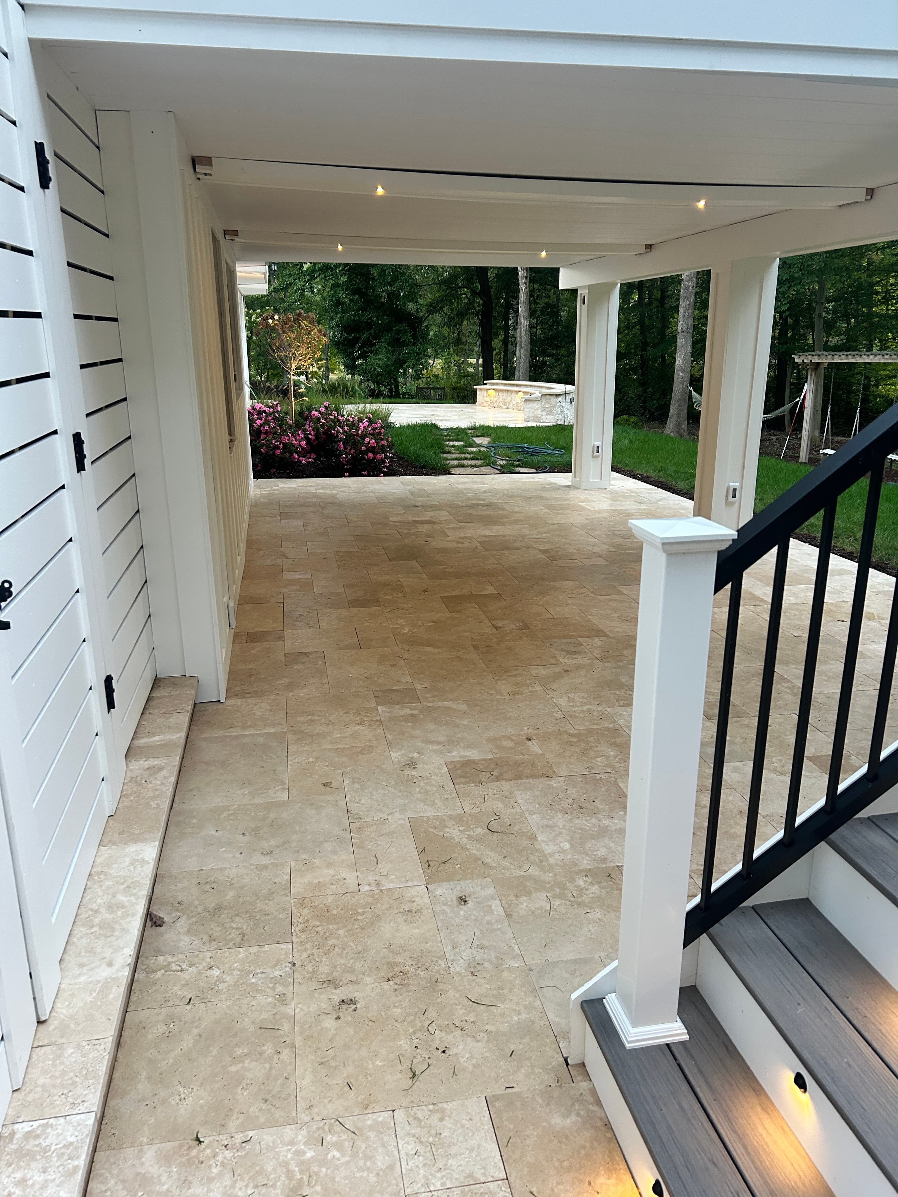 Ivory Travertine Tumbled French Pattern 3cm Paver installed on a covered patio. The natural beige tones and tumbled texture enhance a modern outdoor space with white siding, black railing, and lush greenery, offering a premium hardscape solution by STO...