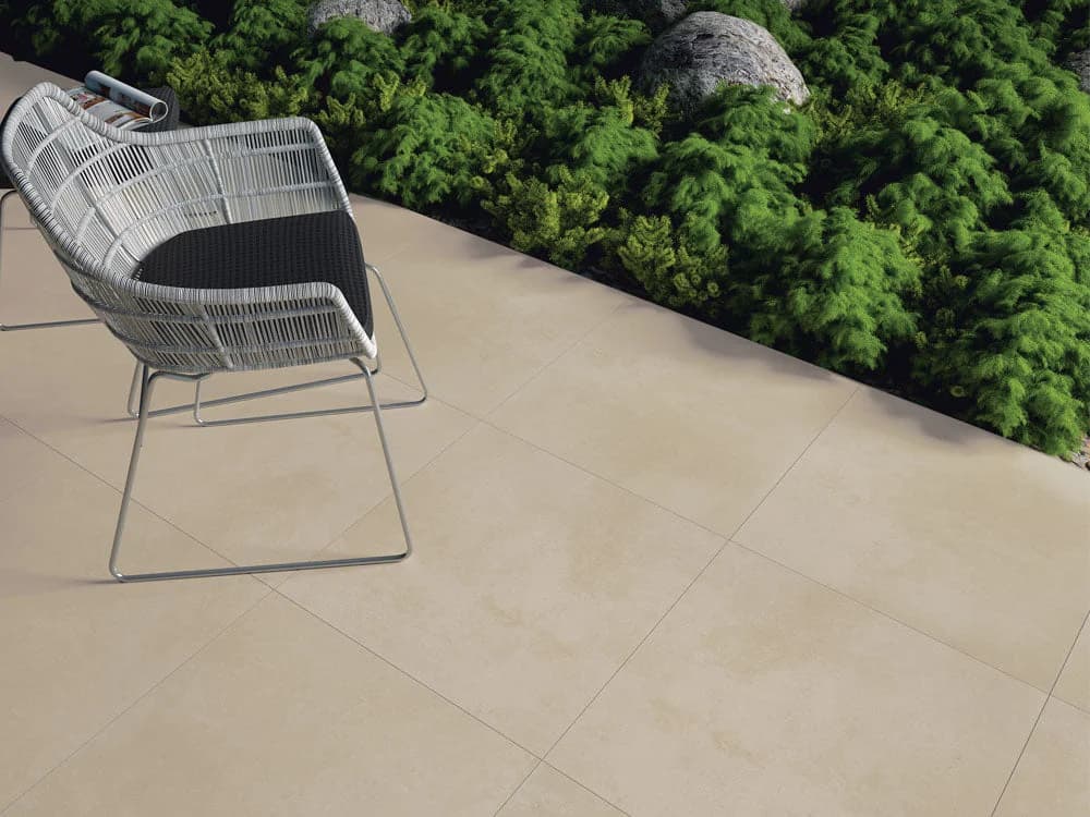 Luna Sand Matte 24x24x2cm Rectified Porcelain Paver installed as patio flooring. The pavers feature a smooth matte finish with subtle sand tones. Paired with modern outdoor wicker chairs, lush green landscaping, and natural stone accents for a contempo...
