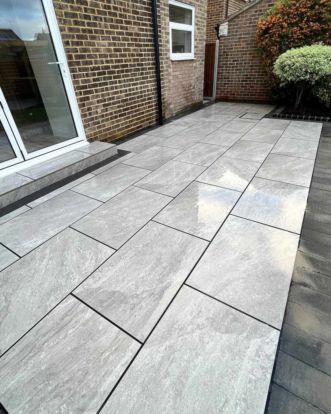 Rasa Gray Matte 24x36x2cm Rectified Porcelain Paver installed as a modern patio. The gray tones and matte finish complement the brick exterior and lush landscaping. Ideal for hardscape projects seeking durable and elegant porcelain solutions.