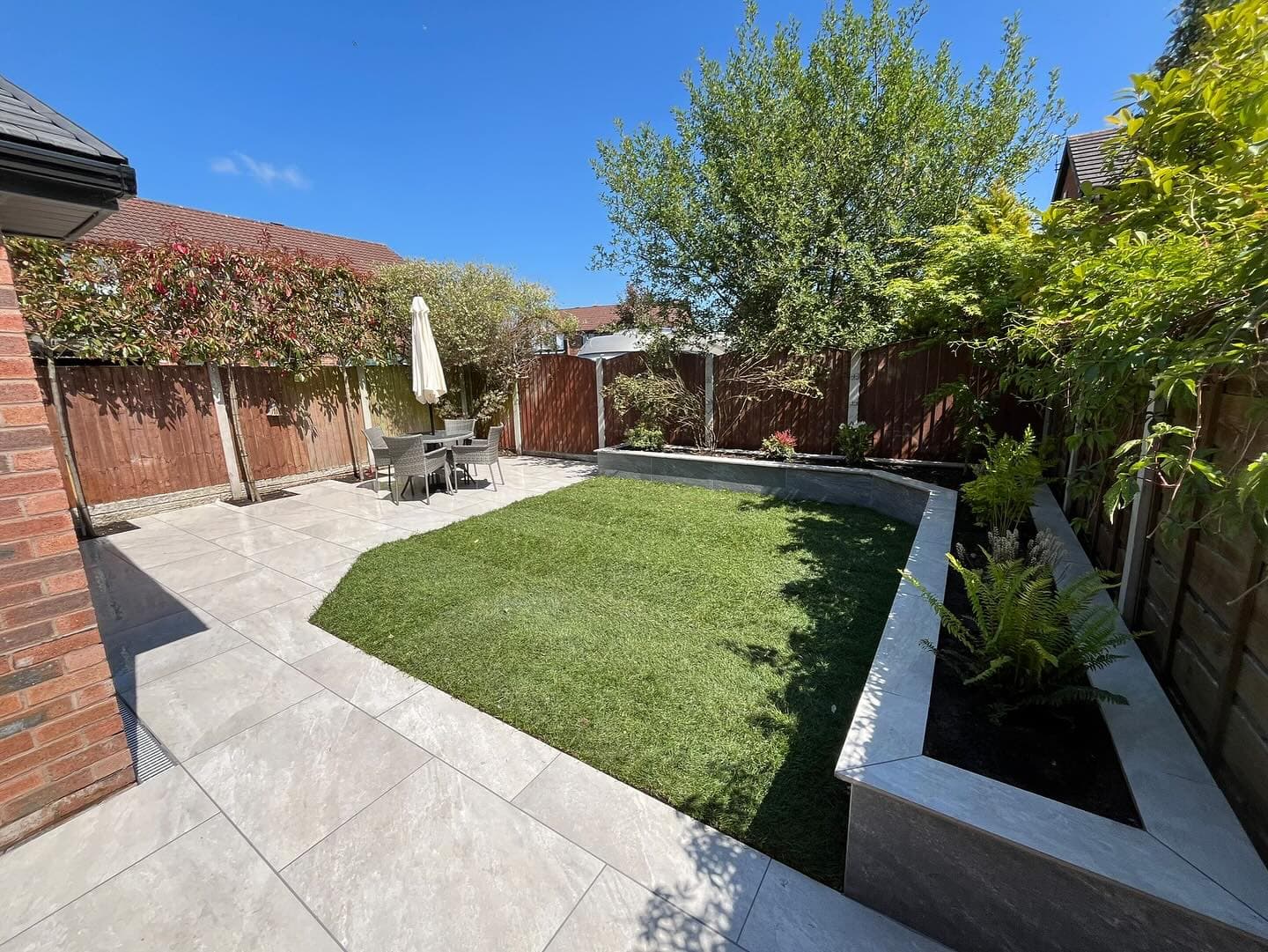 Patio featuring Rasa White Matte 24x24x2cm Rectified Porcelain Pavers by STONELUX. The pavers create a sleek, modern outdoor space with a dining set and lush greenery. Surrounded by wooden fencing, the design complements the vibrant lawn and natural el...