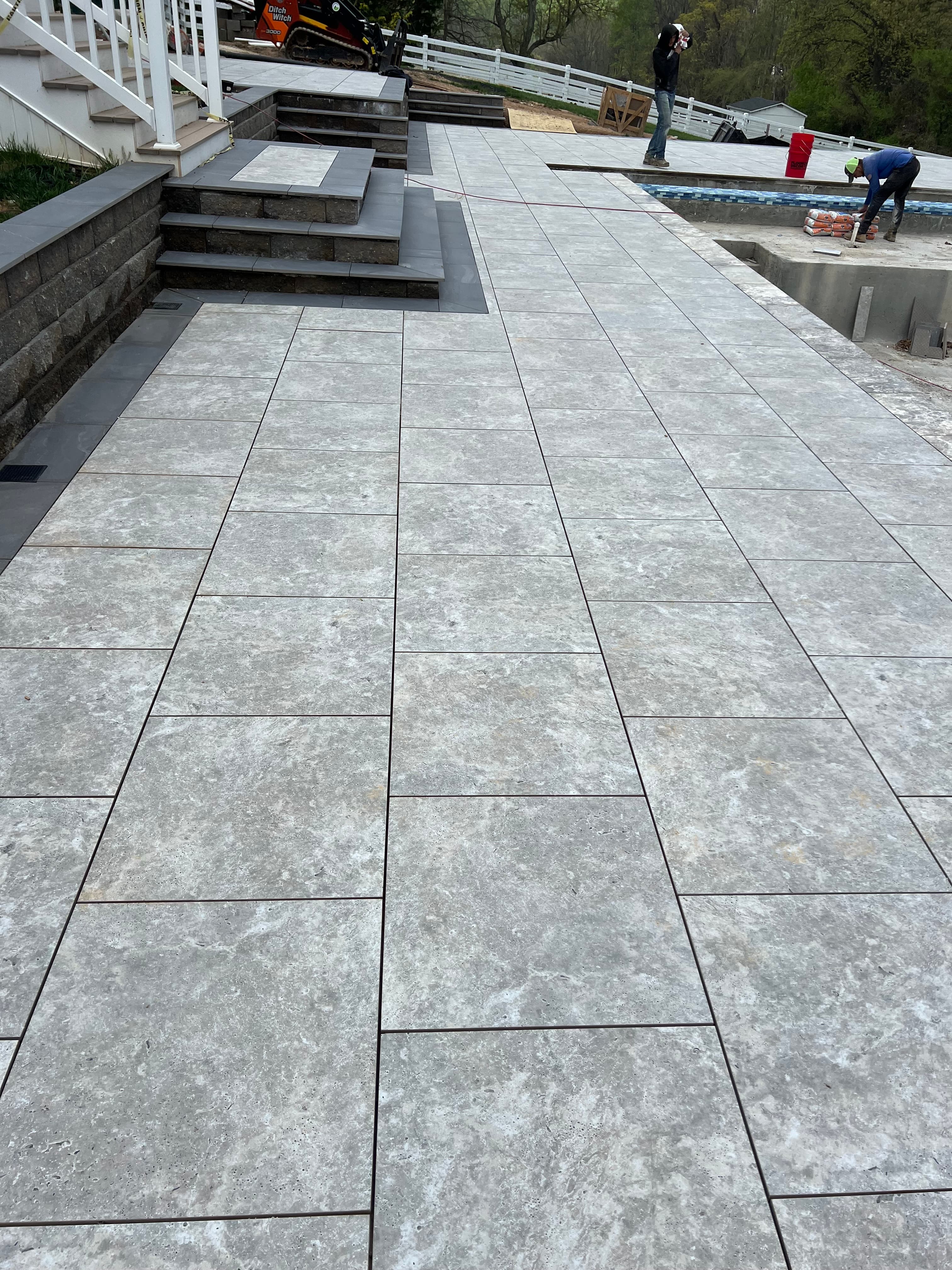 Travertine Silver Matte 24x24x2cm Rectified Porcelain Paver installation on a patio, showcasing a sleek, modern design. The matte finish and natural stone look blend with surrounding landscape, steps, and pool area, ideal for hardscape projects.