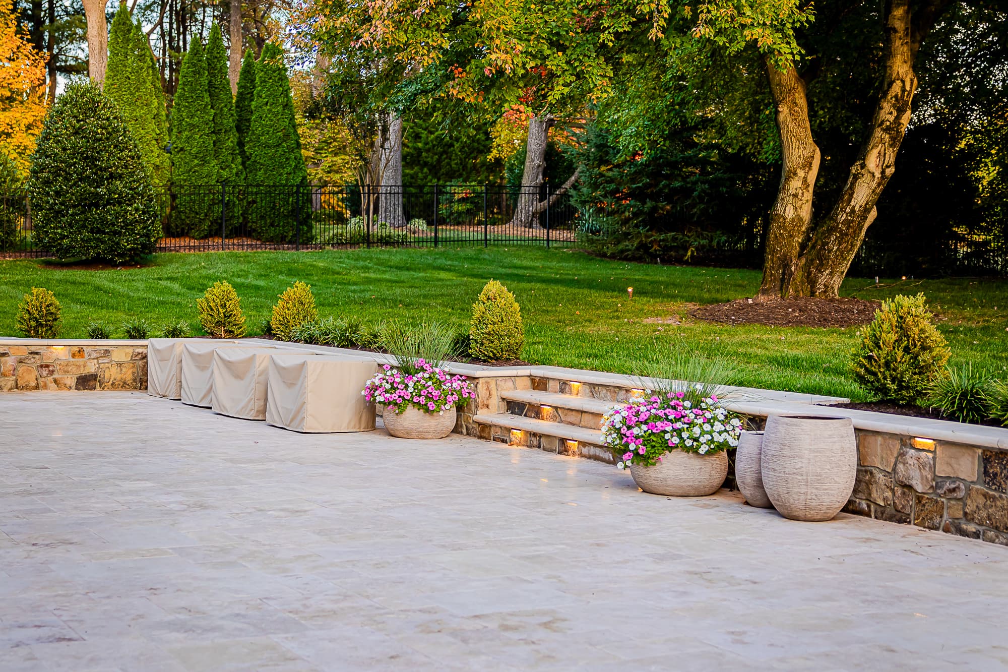 Spacious stone patio, part of the Ivory Twilight: A Travertine Patio Project, features covered seating, colorful planters, softly lit steps, and a landscaped yard with trees and shrubs in the background.