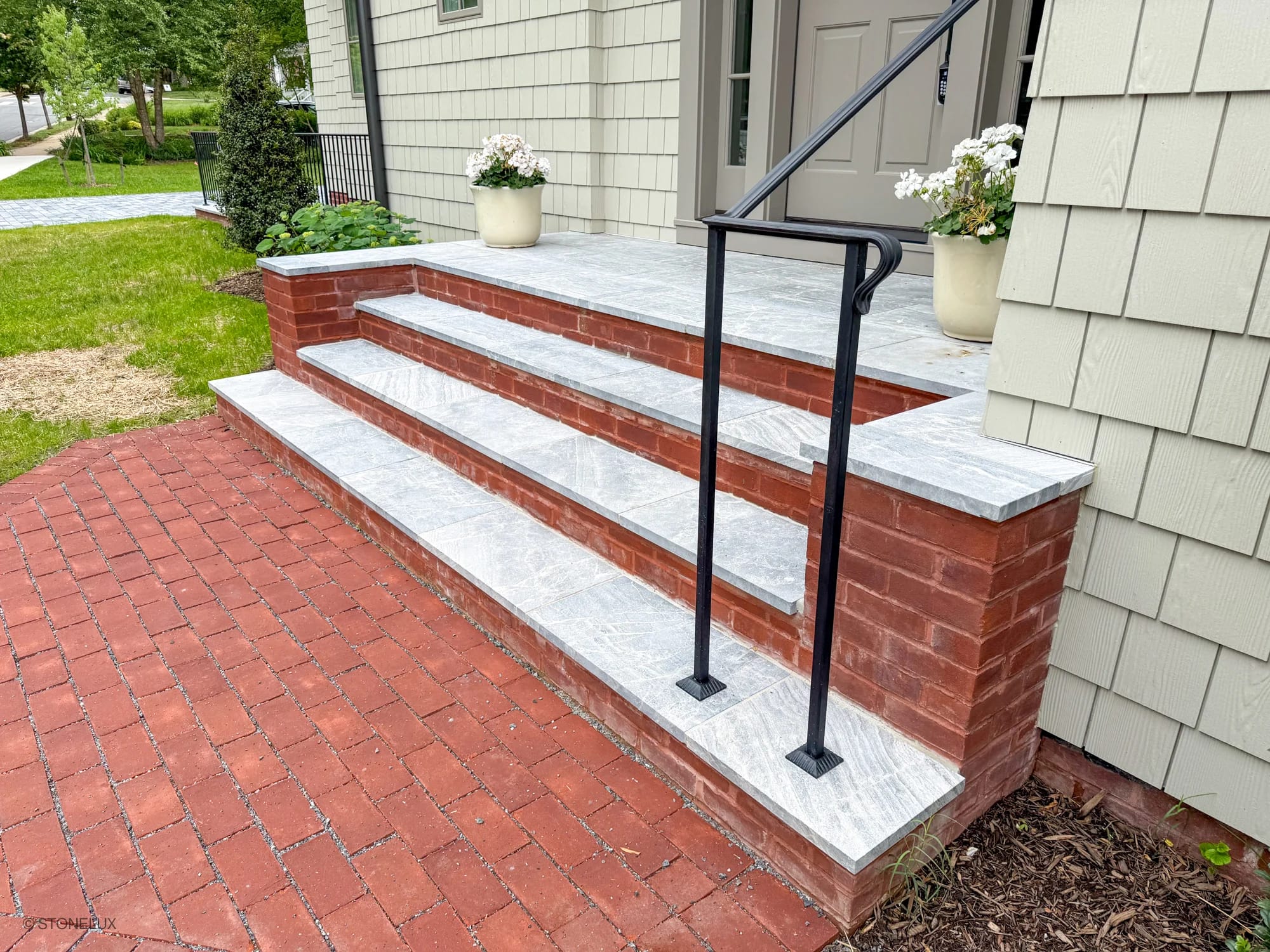 Front entryway showcasing Aegean Grey Marble Sand Blasted 16x24 3cm Paver steps. The elegant marble pavers contrast with red brick, enhancing the traditional architecture. Complemented by green landscaping and a black iron railing for a refined outdoor...