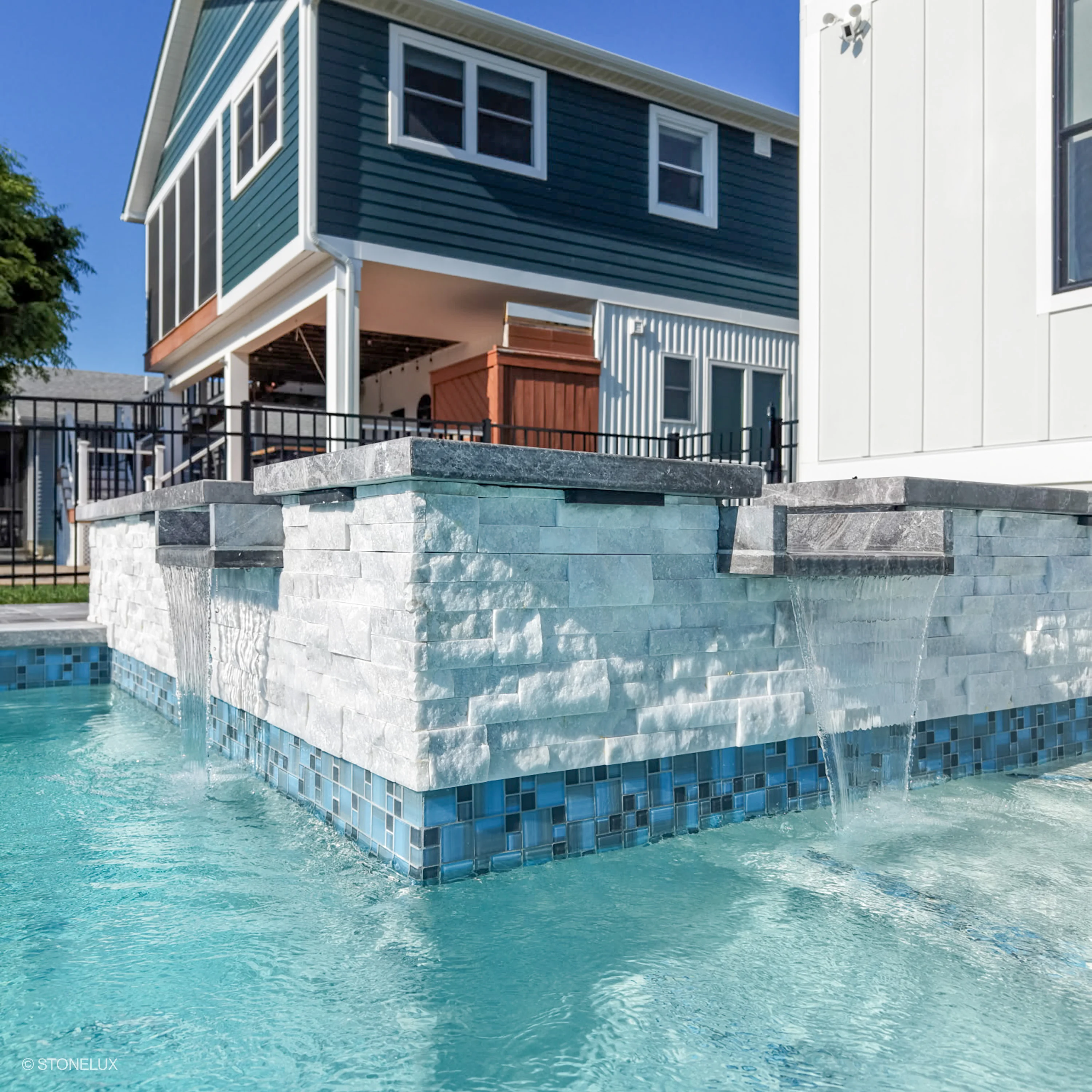 Afyon White Marble Splitface Ledger Panel - 6x24 installed on a modern pool feature wall. The textured marble complements the turquoise water and blue mosaic tiles, enhancing the contemporary outdoor design against a backdrop of a stylish, two-story ho...
