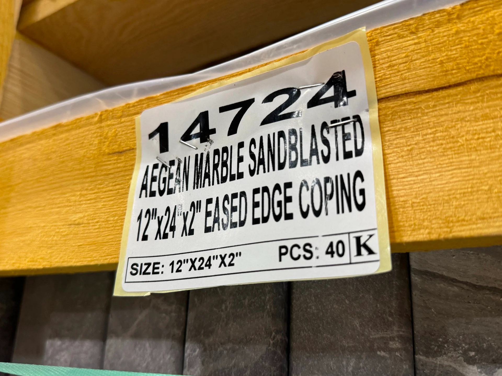 A close-up photo of a warehouse pallet label reading “Serial 14724 Aegean Marble Sandblasted 12"x24"x2" Eased Edge Coping.” The label indicates 40 pieces per crate and is stapled to a wooden crate containing neatly stacked grey marble copings with a sandblasted finish.