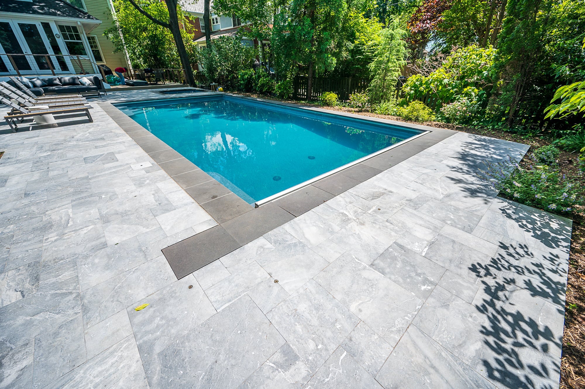 Aegean Grey Marble pool deck installation showcasing elegant, natural stone pavers surrounding a rectangular pool with clear blue water. The marble's subtle veining complements the lush green landscaping and modern outdoor seating, creating a luxurious...