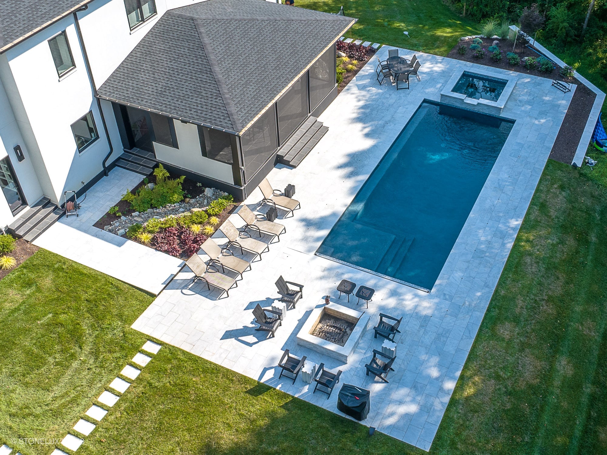 Aerial view of a modern backyard featuring a pool deck and patio crafted with Afyon White Marble from STONELUX. The elegant marble surrounds a rectangular pool with a spa, complemented by sleek outdoor furniture and lush landscaping, creating a luxurio...