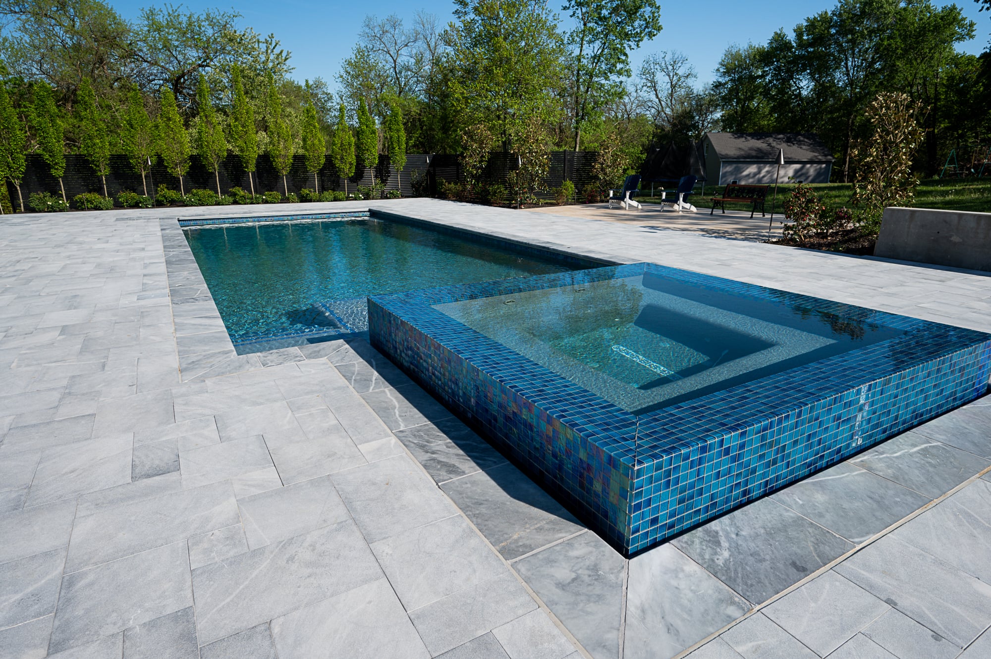 Luxurious outdoor pool area featuring Arctic Grey Marble pavers by STONELUX. The natural stone complements the vibrant blue pool tiles, surrounded by lush greenery. Ideal for modern hardscape projects and pool renovations.
