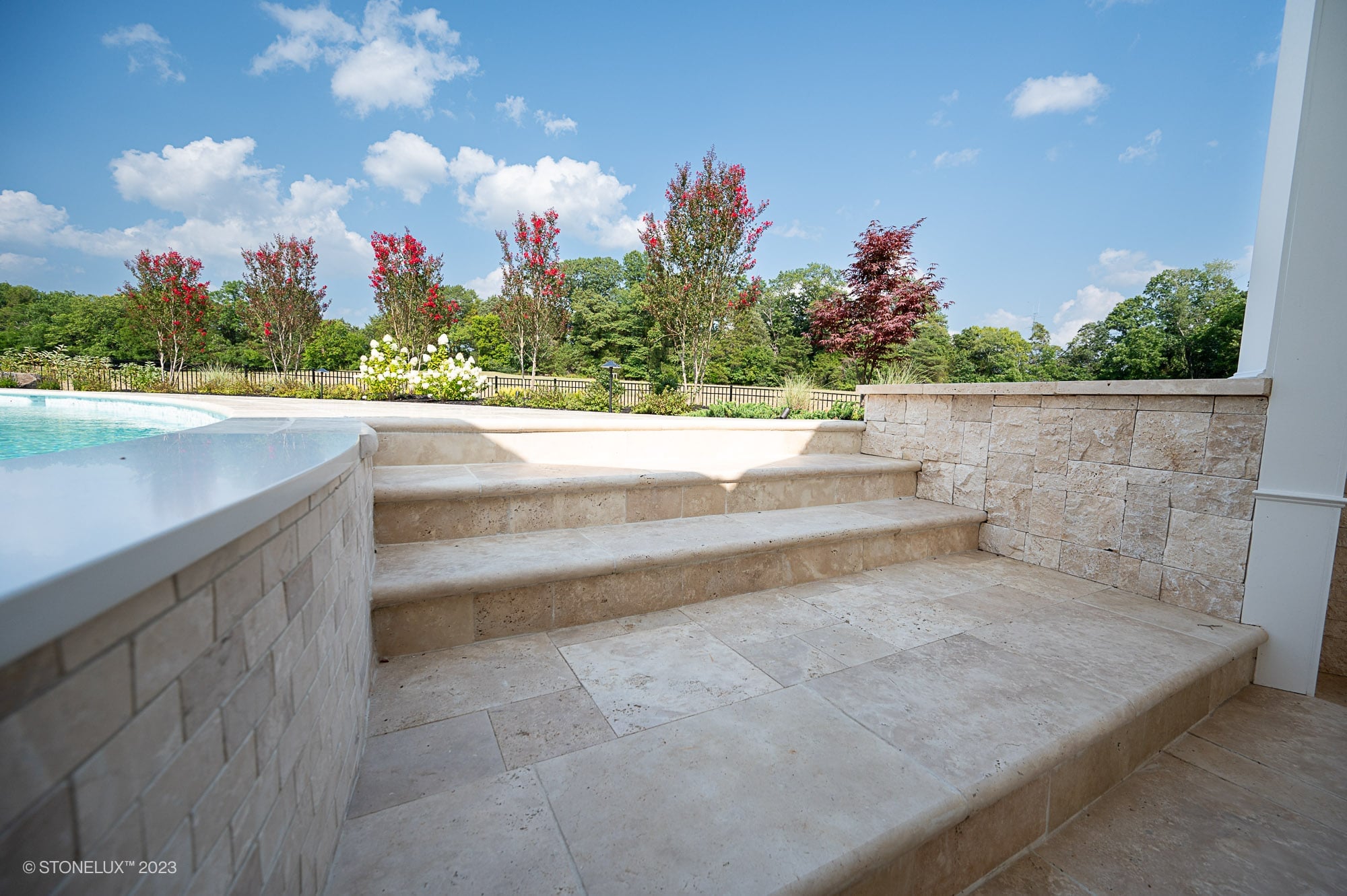 Ivory Travertine natural stone steps and pool coping create a seamless transition between the pool area and garden. The light beige tones and subtle veining enhance the outdoor landscape with vibrant red and green foliage, offering a serene, elegant look.
