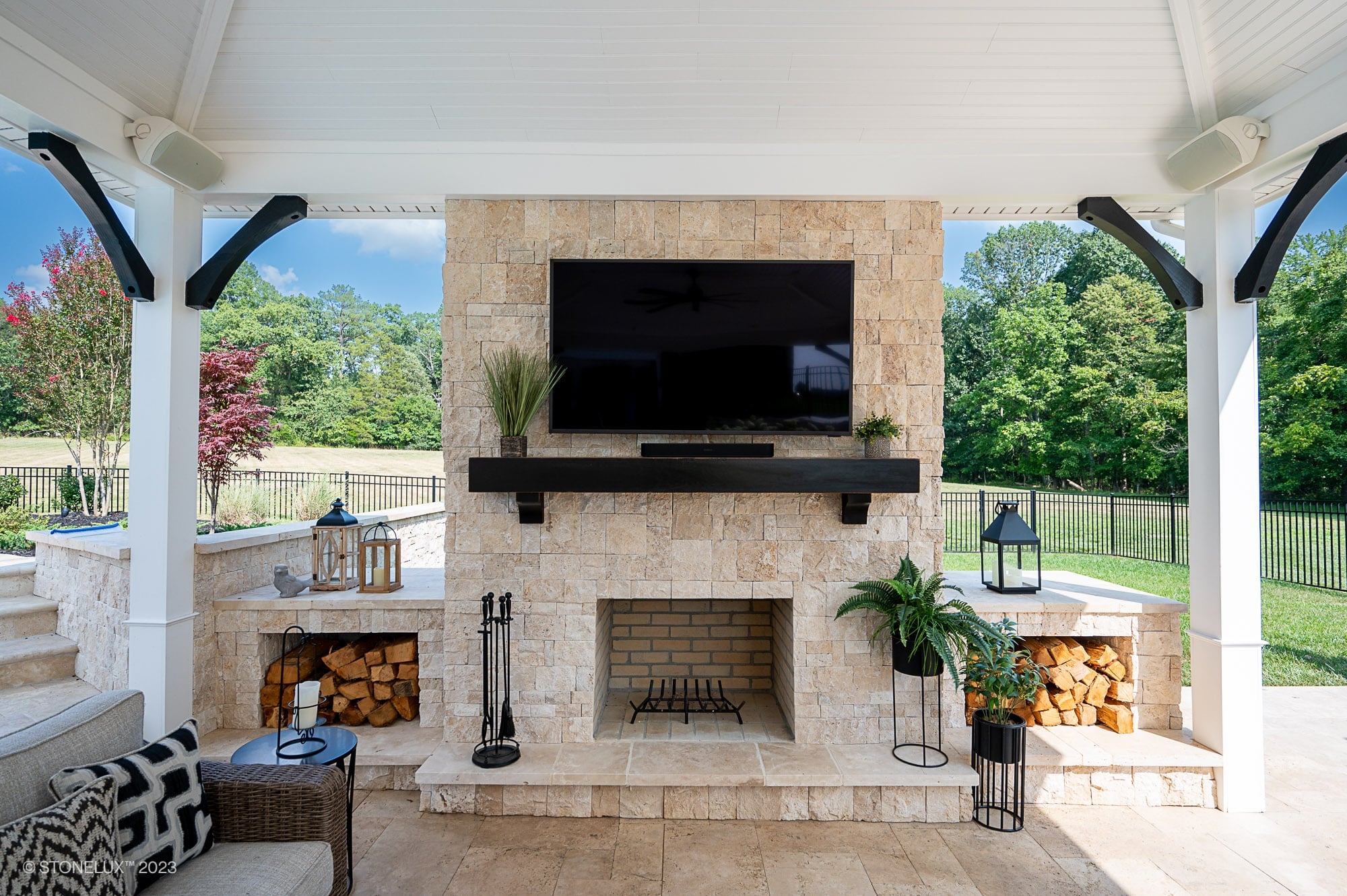 Outdoor living space featuring STONELUX Ivory Travertine natural stone used for a fireplace surround and flooring. The elegant patio includes modern black accents, a TV, and lush greenery, creating a harmonious blend of style and nature. Ideal for luxu...
