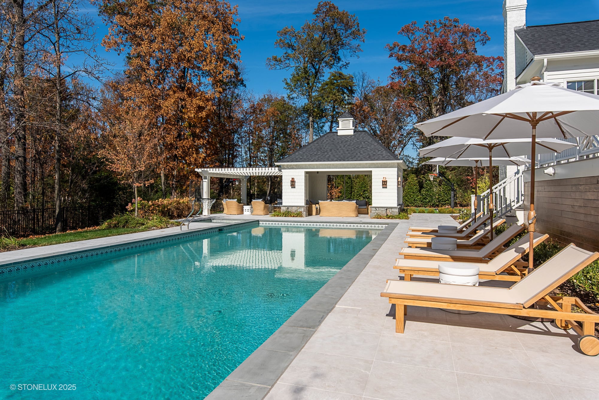 Luxurious poolside featuring Pera Cream Marble pavers by STONELUX. Elegant natural stone surrounds a sparkling blue pool, complemented by chic loungers and umbrellas. The scene includes a stylish pool house and pergola, enhancing the modern outdoor oasis.