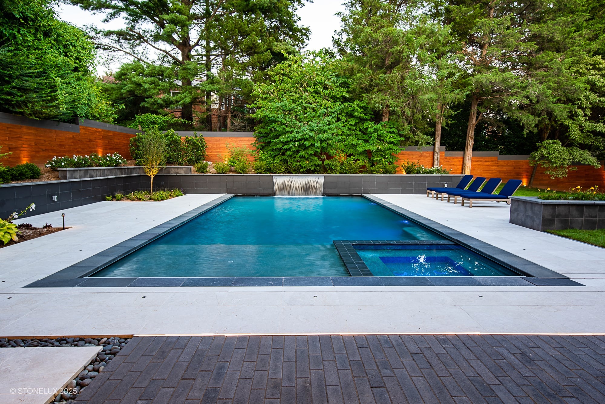 Luxurious modern pool design featuring Pera Cream Marble pool deck by STONELUX. Highlighting elegant marble coping, blue-tinted water, sleek black tiles, and lush greenery. Perfect for premium hardscape projects and outdoor renovations.