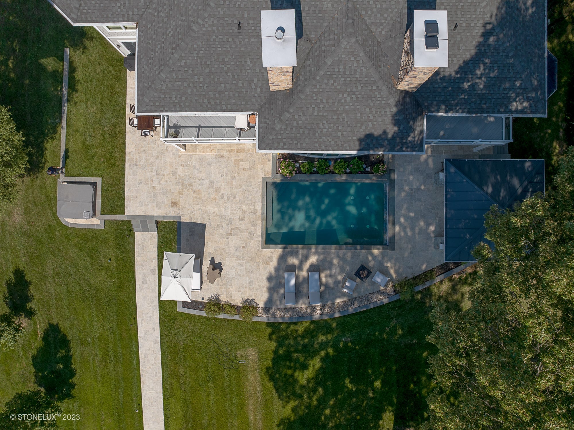 Aerial view of a luxurious outdoor space featuring Philly Travertine natural stone as pool deck and patio pavers. The design includes a rectangular pool with clear blue water, modern outdoor furniture, lush green lawn, and elegant landscaping, enhancin...