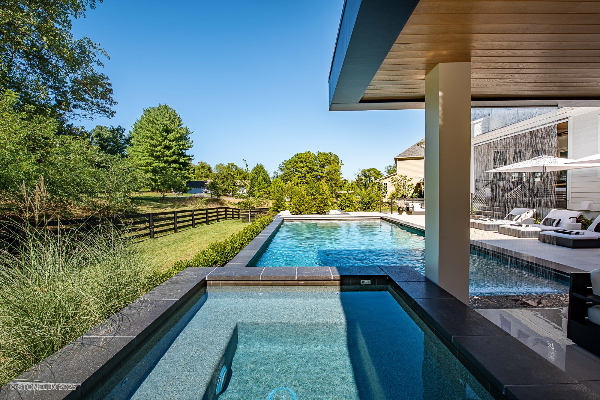 Luxury outdoor pool featuring Black Basalt natural stone coping from STONELUX. Modern design with clear blue water, lush landscaping, and elegant poolside furniture under a sleek patio cover, ideal for upscale hardscape and pool renovations.