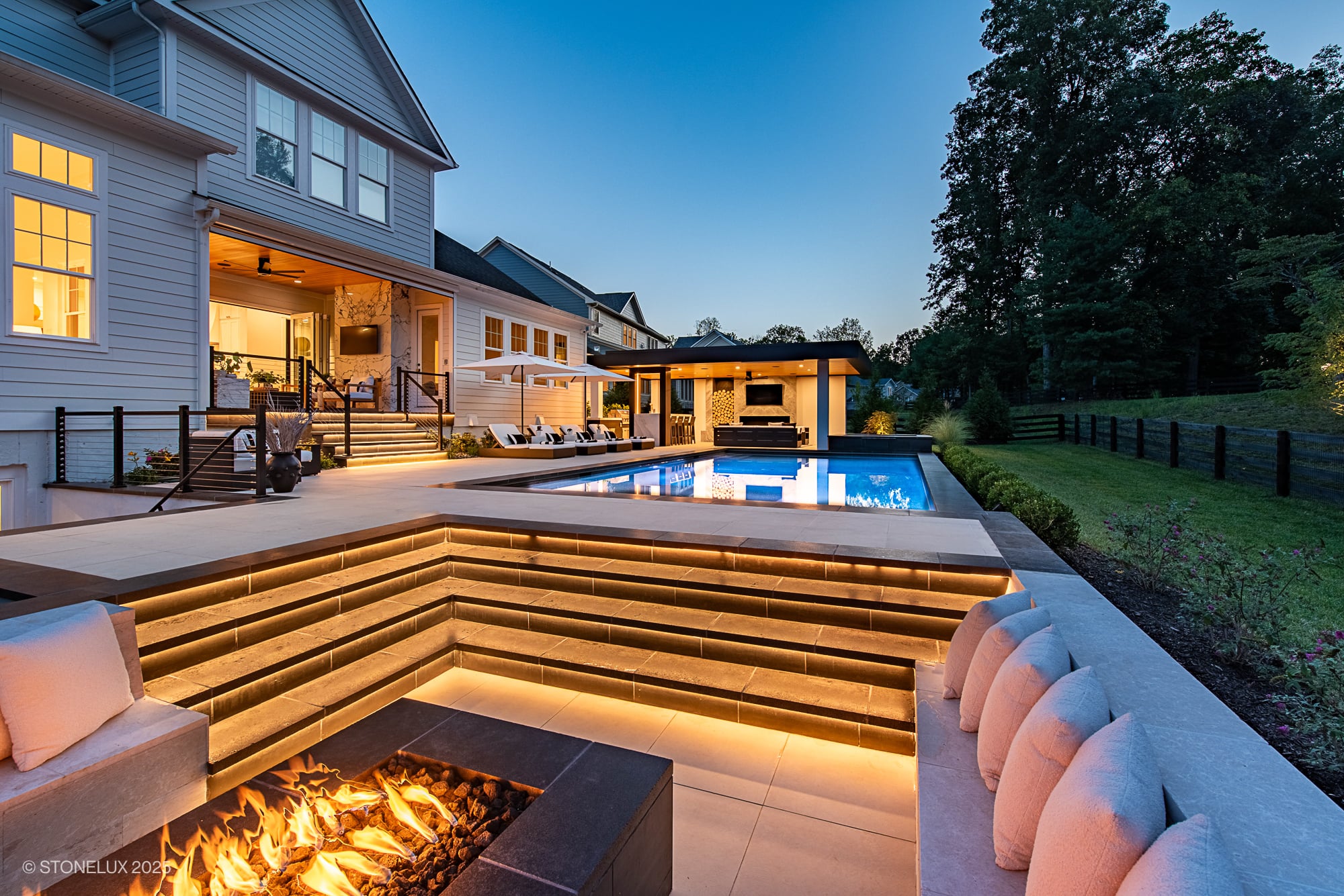 Luxurious modern backyard with pool featuring STONELUX Black Basalt natural stone. The basalt is used for elegant pool coping and steps, accented by warm lighting. The scene includes a fire pit, contemporary outdoor furniture, and lush greenery, creati...