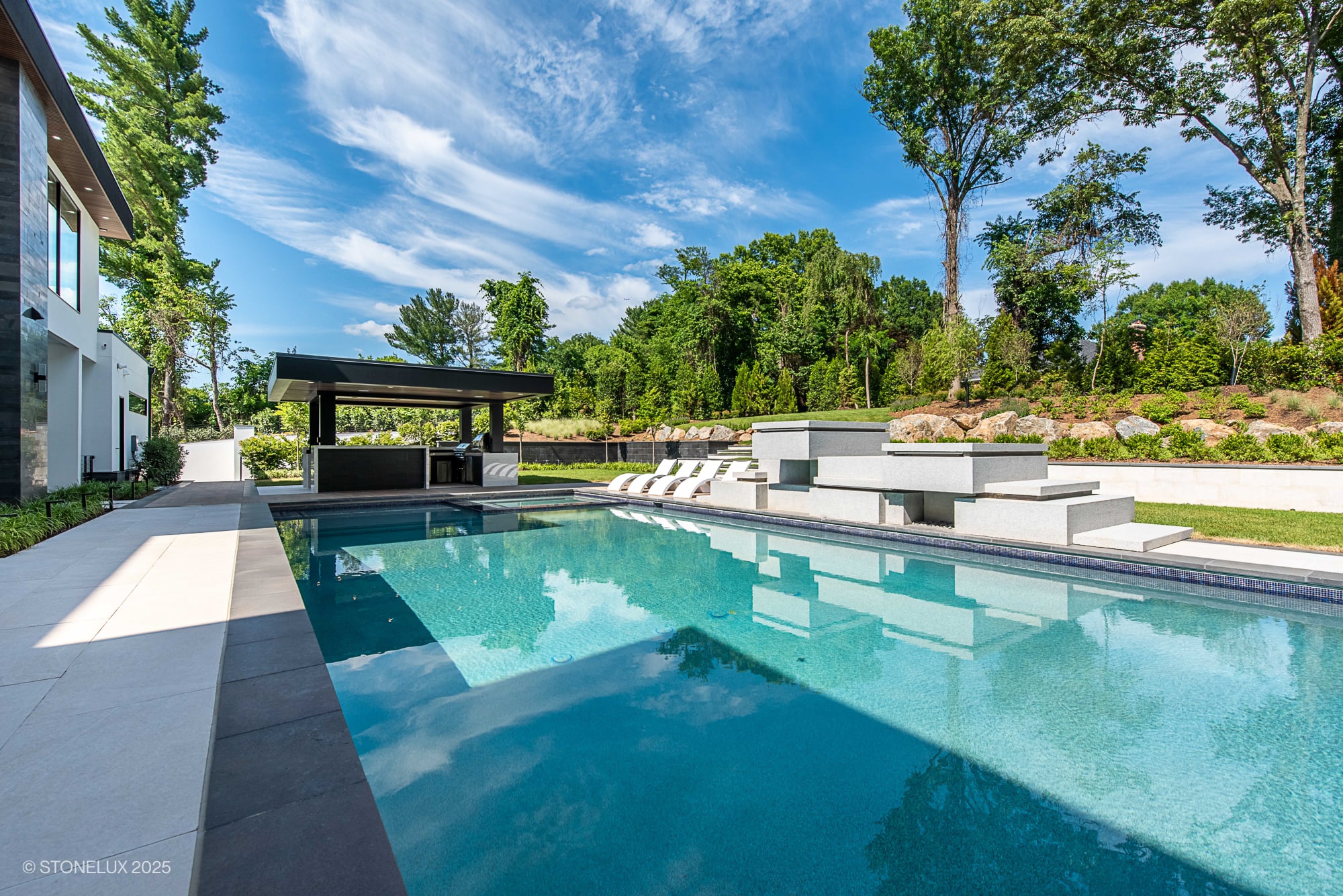 Modern poolside installation featuring STONELUX Porcelain Paver in natural stone. The sleek pool deck complements the clear blue water, surrounded by lush landscaping and a contemporary outdoor kitchen. Ideal for luxury hardscape projects.
