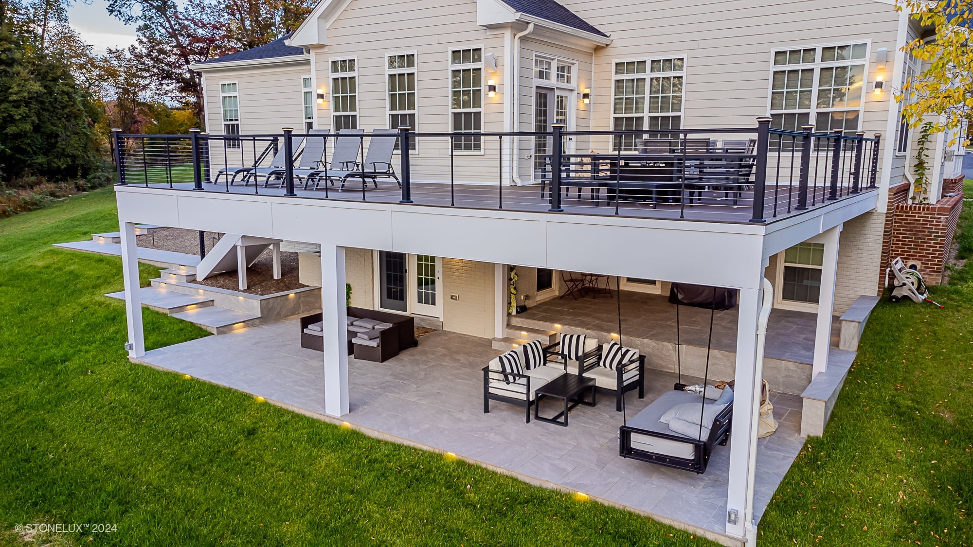 Modern two-story patio installation featuring STONELUX Porcelain Pavers in a natural stone finish. Upper deck with lounge chairs and dining area, lower patio with outdoor seating. Surrounded by lush greenery, contemporary architecture, and ambient ligh...