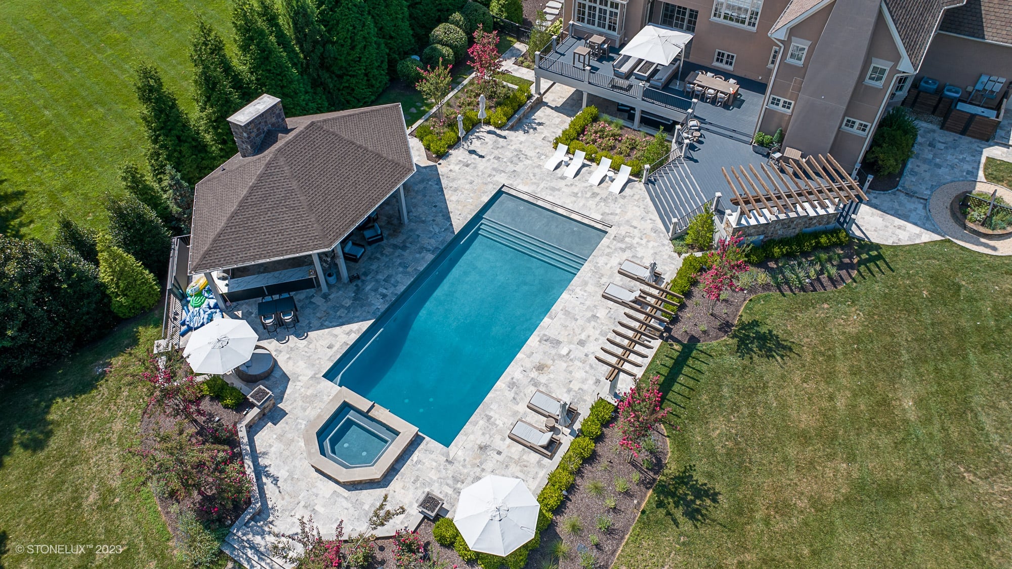 Aerial view of a luxurious pool area featuring Silver Travertine pavers by STONELUX. The natural stone pool deck surrounds a rectangular pool and spa, complemented by lush landscaping, a pergola, and modern patio furniture, creating an elegant outdoor ...