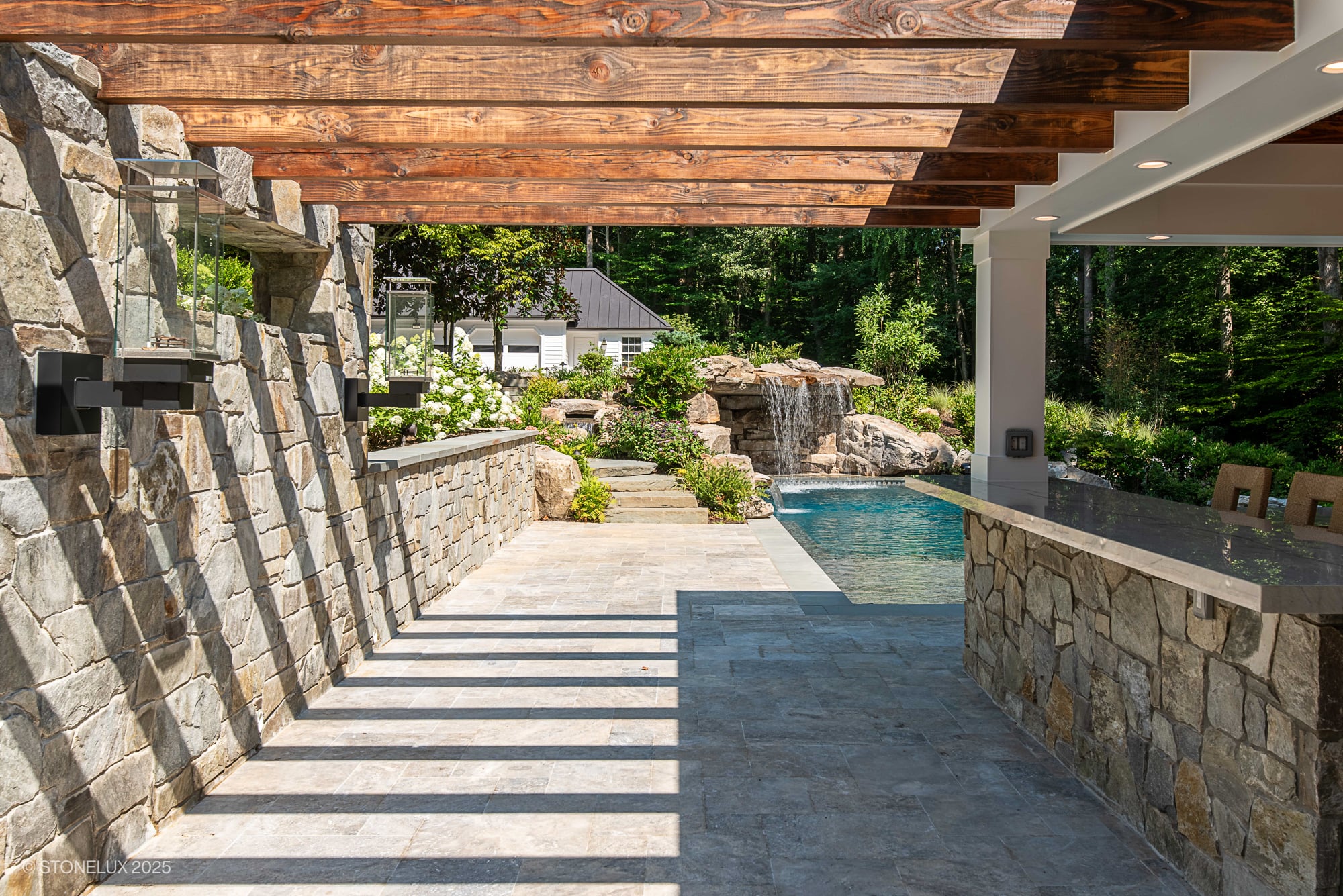 Outdoor oasis featuring STONELUX Silver Travertine pavers in a luxury patio setting. The natural stone complements rustic wood beams, a stone wall, and a sparkling pool with a waterfall, surrounded by lush greenery and modern outdoor furniture.