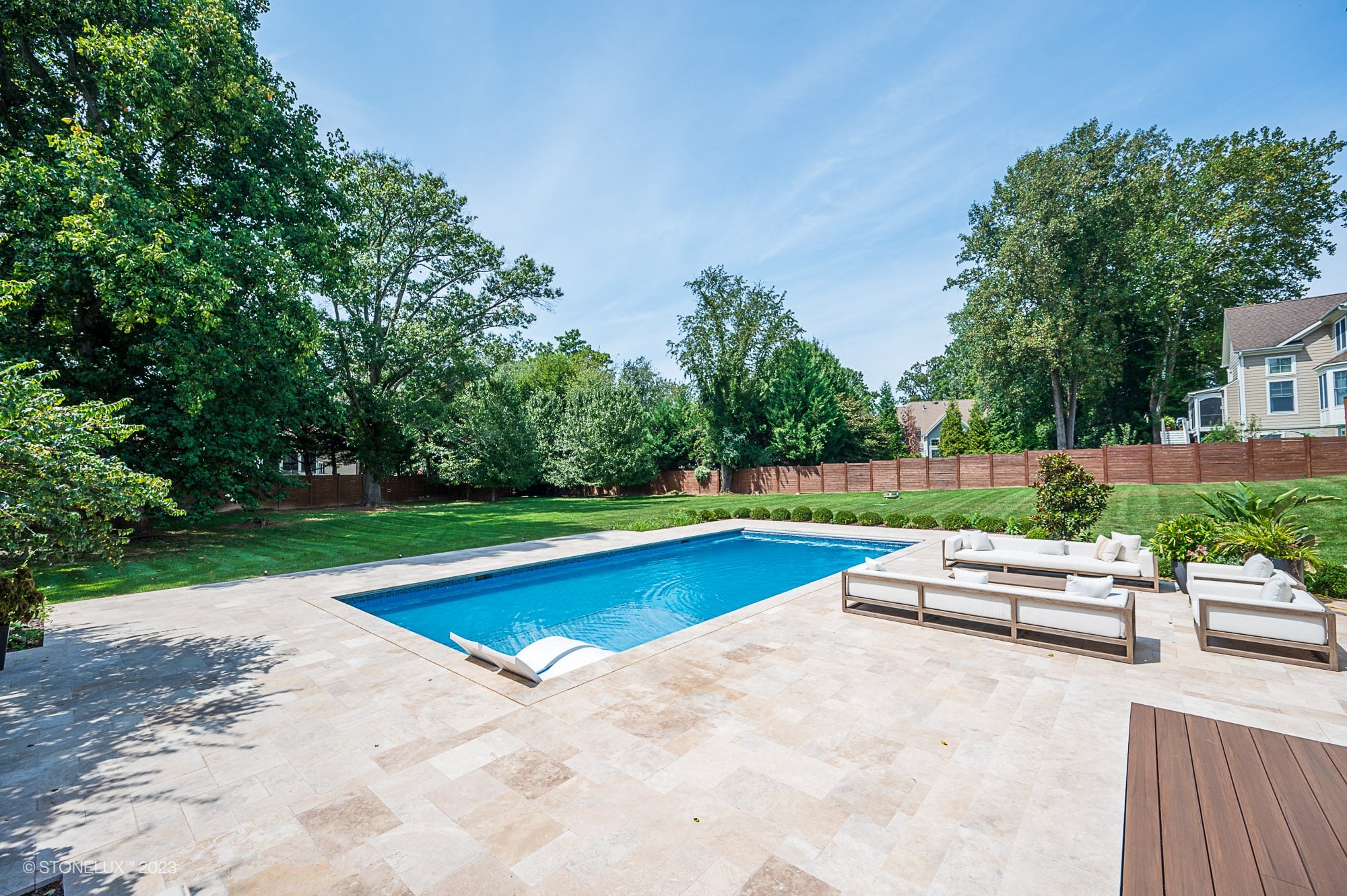 Luxurious backyard installation featuring Walnut Travertine natural stone pavers by STONELUX surrounding a sparkling blue pool. Elegant patio with modern outdoor seating, lush green landscaping, and a wooden privacy fence create a tranquil, stylish ret...