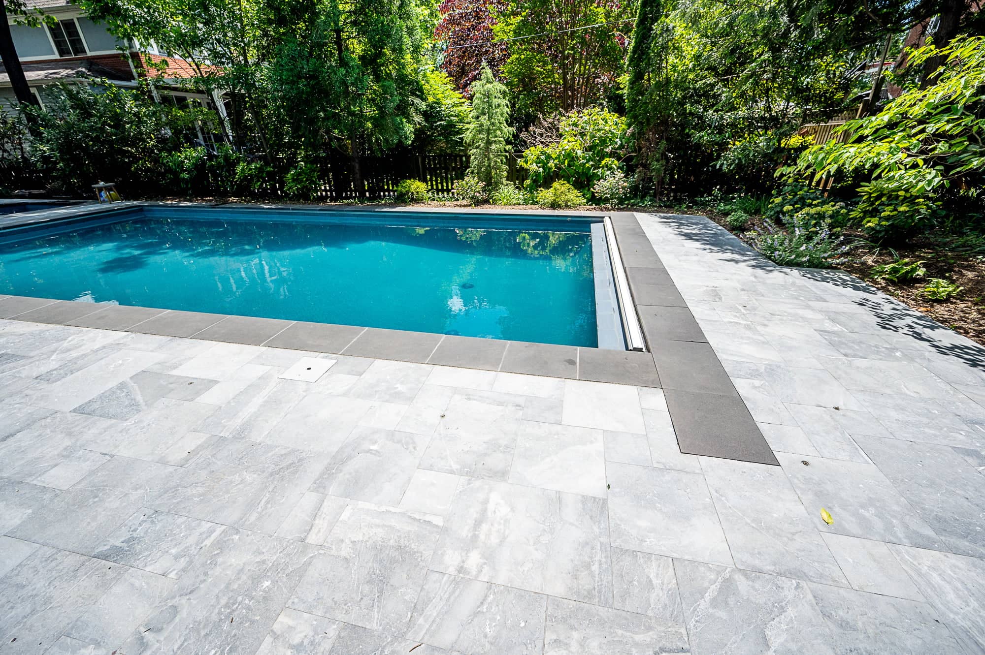 Aegean Grey Marble pool deck featuring elegant natural stone with subtle veining, complementing the clear blue pool water. Surrounded by lush greenery, this modern outdoor space showcases marble pavers with dark coping, creating a luxurious, serene atm...