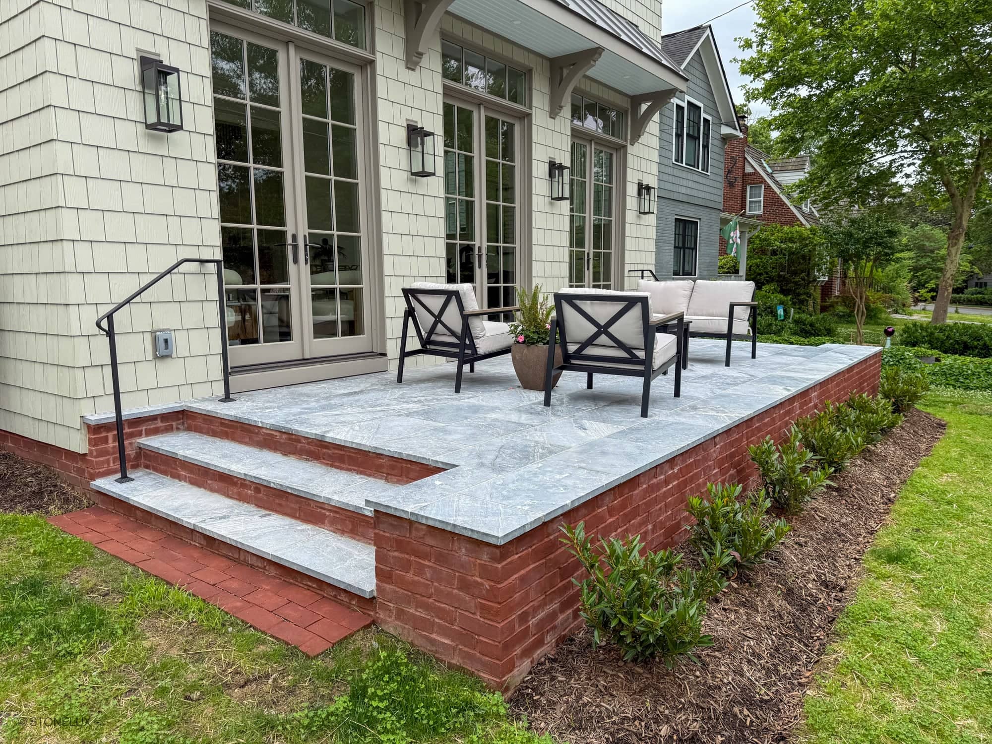 Outdoor patio featuring Aegean Grey Marble pavers by STONELUX, installed as a sleek surface atop red brick steps. The modern seating area, surrounded by lush greenery and classic architecture, showcases the natural stone's subtle veining and elegance.