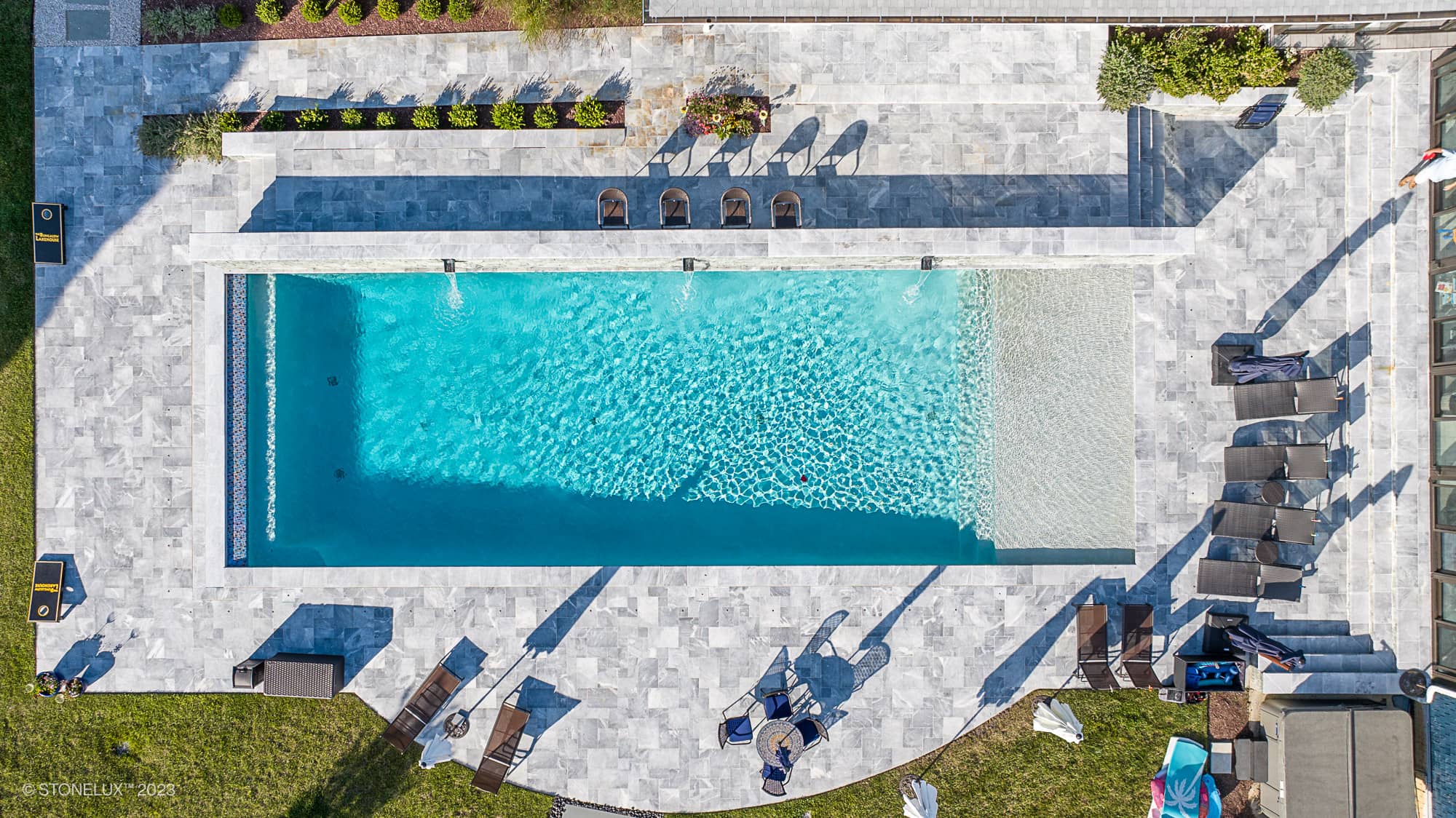 Aerial view of a modern pool deck featuring Arctic Grey Marble by STONELUX. The natural stone pavers surround a vibrant blue pool, complemented by elegant patio furniture and lush landscaping, creating a luxurious outdoor oasis.
