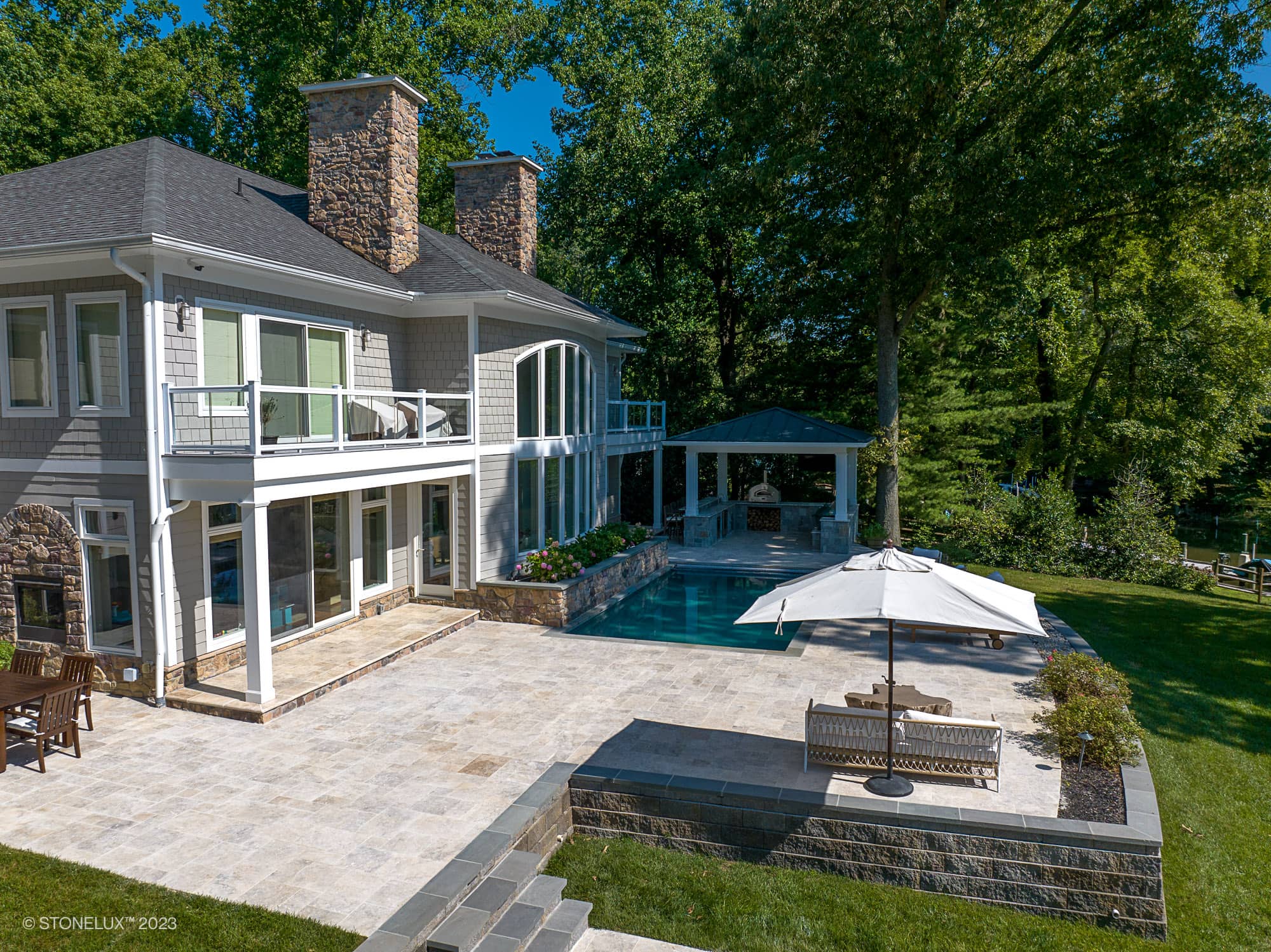 Luxurious outdoor patio featuring Philly Travertine natural stone pavers by STONELUX. Installed around a modern pool with lush landscaping, a gazebo, and elegant patio furniture. Perfect for upscale hardscape projects and pool renovations.