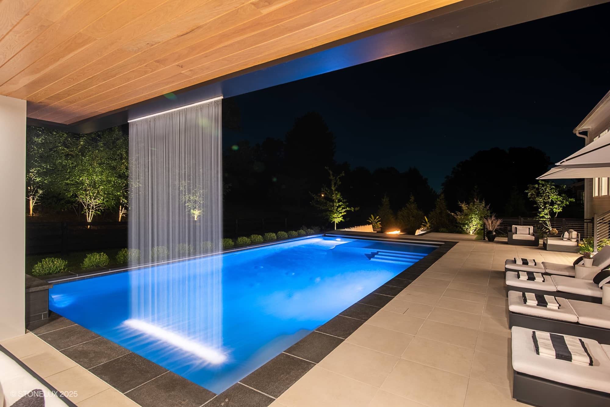 Luxurious poolside installation featuring Stonelux Black Basalt natural stone as pool coping. The modern design includes a striking waterfall feature, ambient lighting, and elegant patio seating, complemented by lush landscaping and a serene blue pool.