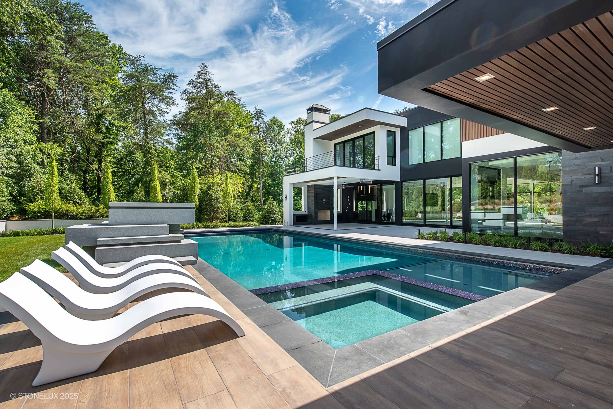 Modern poolside scene featuring STONELUX Black Basalt natural stone as sleek pool coping, surrounded by lush greenery. Contemporary architecture complements the design with clear blue pool water and stylish white lounge chairs on a wooden deck.