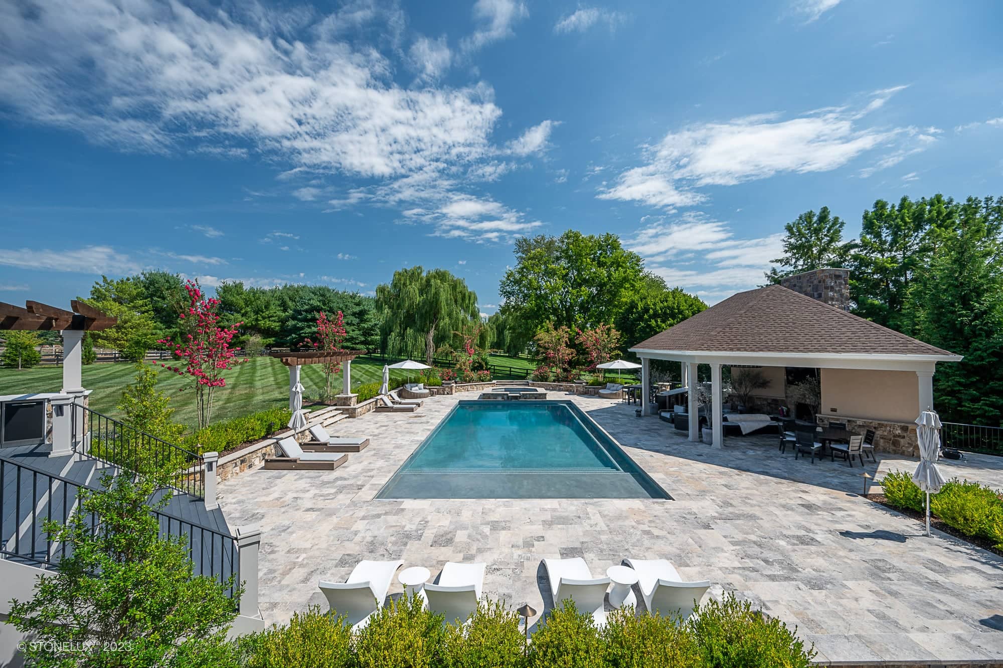 Luxurious outdoor pool area featuring Silver Travertine natural stone as pool deck pavers. Surrounded by lush landscaping, modern lounge chairs, and a stylish cabana, creating a serene setting. Perfect for hardscape projects and premium pool renovations.