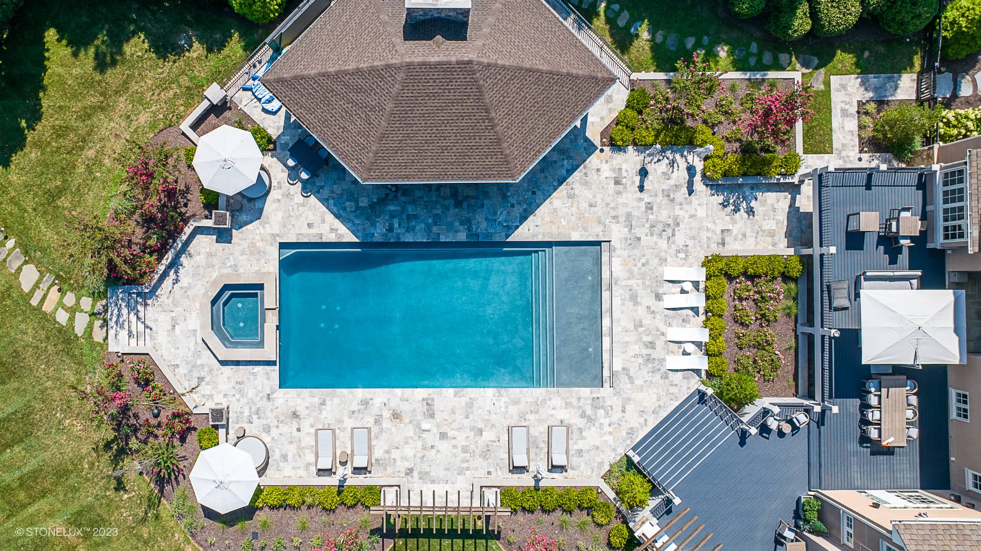 Aerial view of a luxurious pool area featuring Silver Travertine natural stone pavers. The installation includes a pool deck and patio, surrounded by lush landscaping, white lounge chairs, and umbrellas. The elegant design blends seamlessly with the mo...