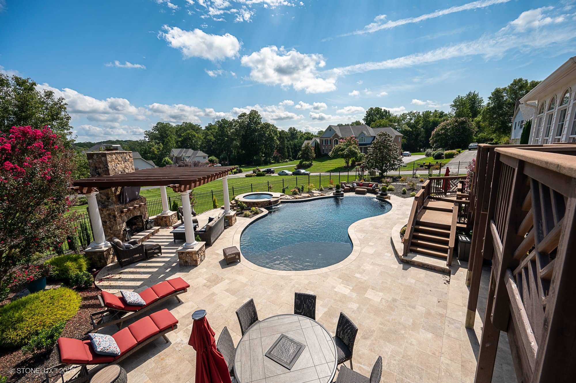 Luxurious outdoor pool area featuring Walnut Travertine natural stone pavers by STONELUX. The pool deck showcases elegant travertine tiles, complemented by a stone fireplace, red loungers, and lush green landscaping, creating a serene and stylish retreat.