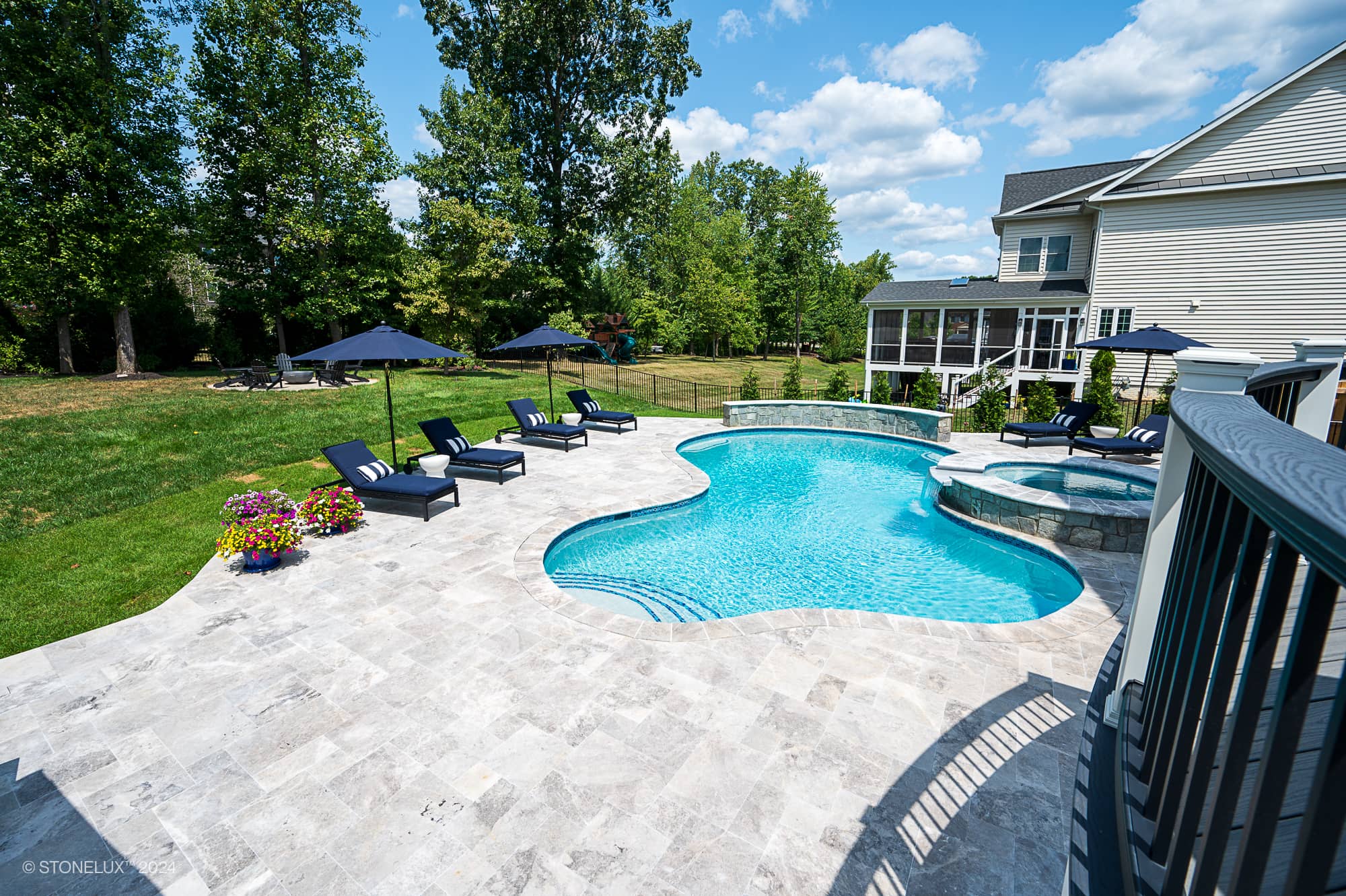 Luxurious backyard pool area featuring Tundra Grey Marble pavers from STONELUX. The elegant natural stone surrounds a curvy pool with crystal blue water, complemented by navy loungers, umbrellas, vibrant flowers, and lush green landscaping. Perfect for...