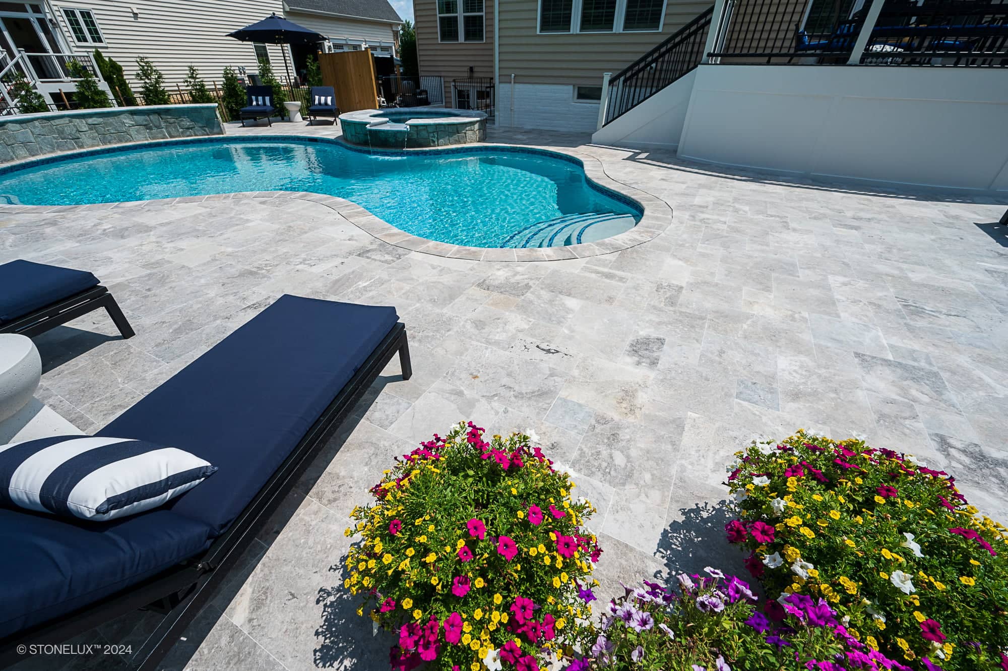 Outdoor pool area featuring Tundra Grey Marble pavers by STONELUX. The elegant natural stone enhances the pool deck, surrounded by vibrant flowers, modern navy loungers, and a sparkling blue pool, creating a luxurious and inviting hardscape design.