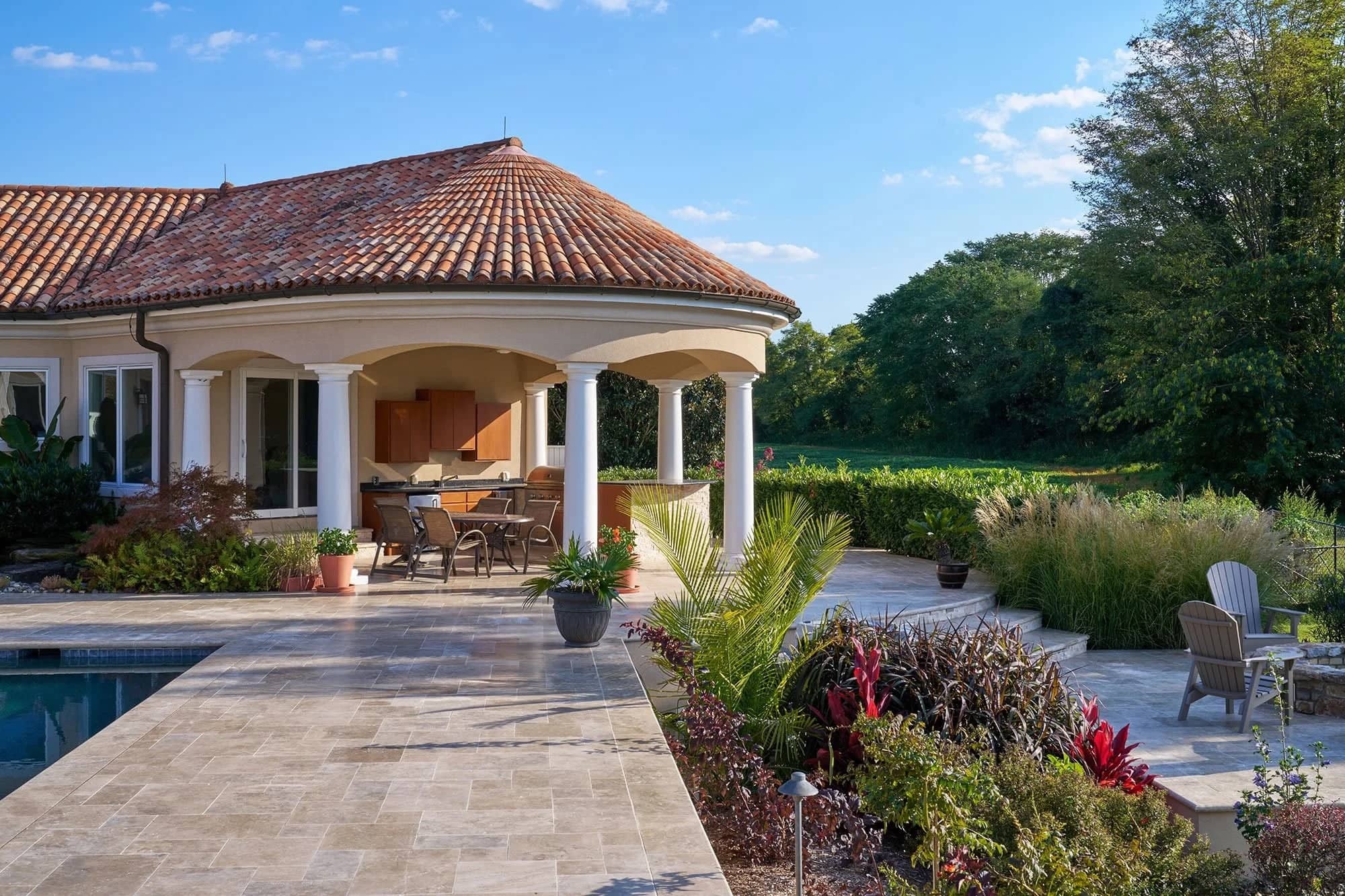 Luxurious outdoor scene featuring Philly Travertine natural stone pavers used in a spacious patio. Surrounded by lush greenery, the elegant setup complements a Mediterranean-style villa with terracotta roof, white columns, and outdoor dining area, idea...
