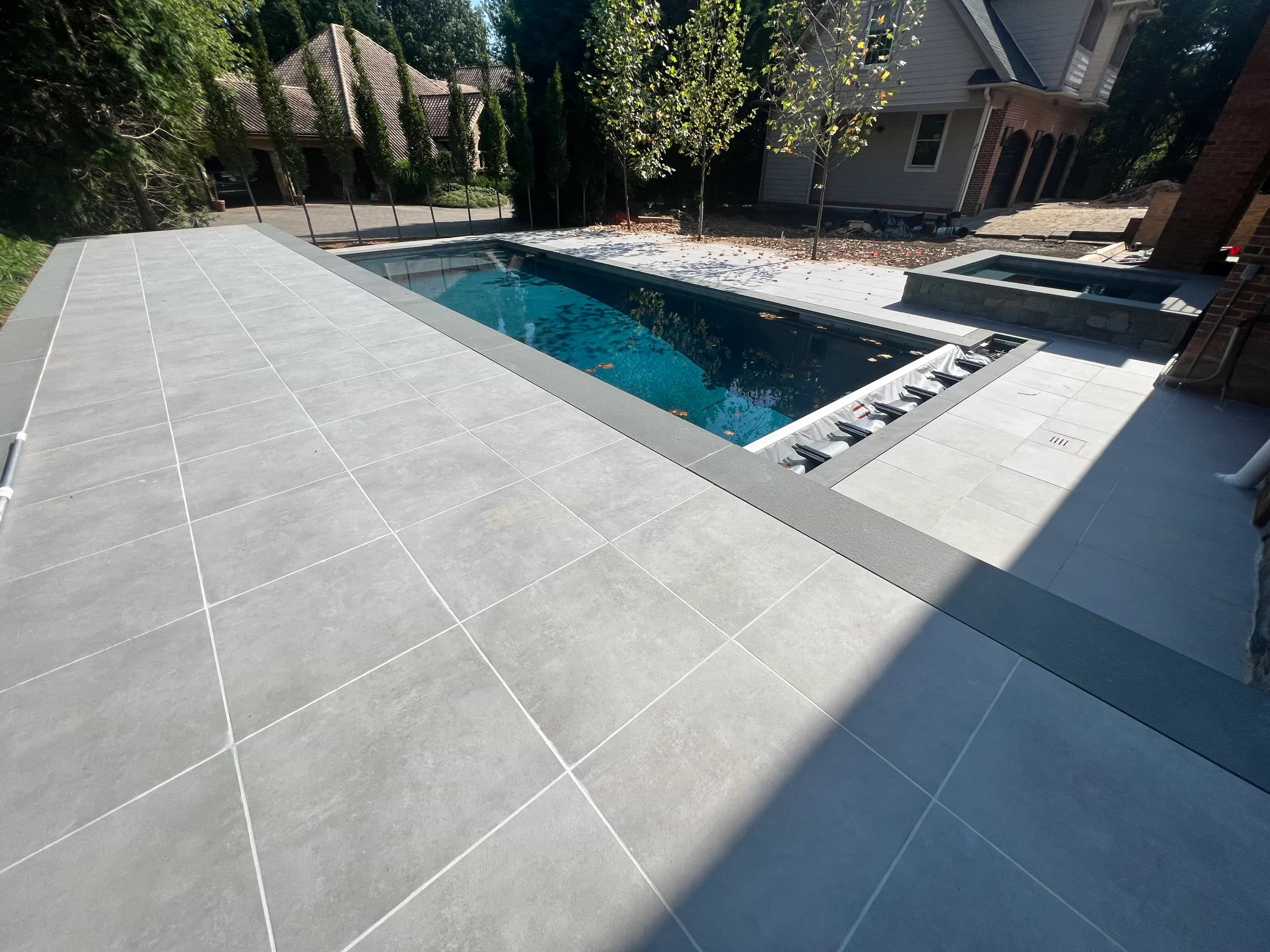 Ark Silver Matte 24x48x2cm Rectified Porcelain Paver installed as a sleek pool deck, enhancing a modern outdoor space. The light gray matte finish complements the deep blue pool water, surrounded by lush greenery and a traditional house, creating a ser...