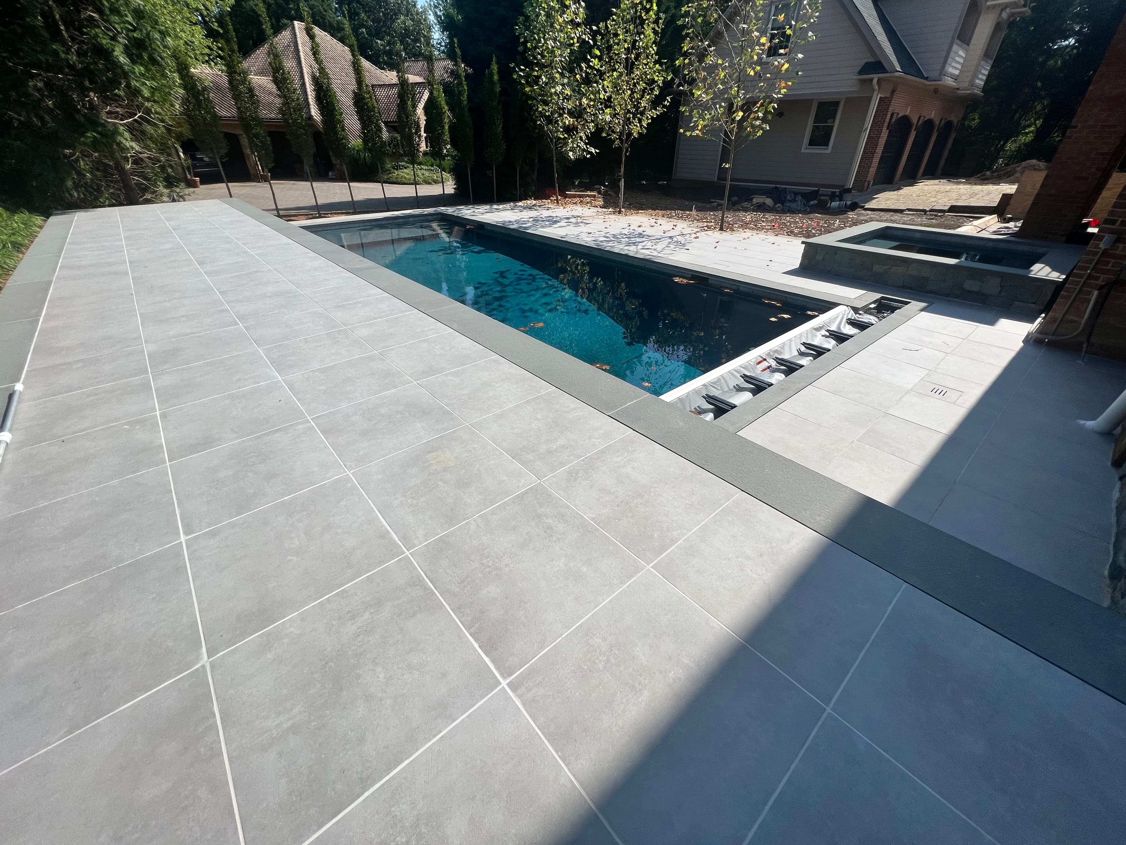 Ark Silver Matte 24x48x2cm Rectified Porcelain Paver installed as a sleek pool deck, enhancing a modern outdoor space. The light gray matte finish complements the deep blue pool water, surrounded by lush greenery and a traditional house, creating a ser...