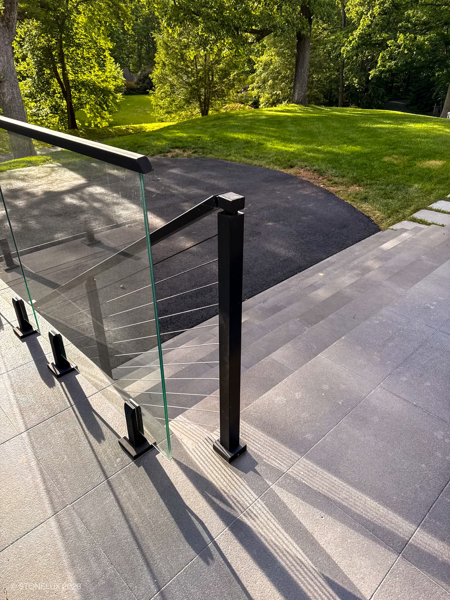 Outdoor front entrance steps featuring STONELUX Black Basalt Tumbled+Brushed 8x16 3cm Pavers, used as elegant steps leading to a lush green lawn. Modern glass railing complements the sleek stone surface, enhancing the contemporary landscape design.