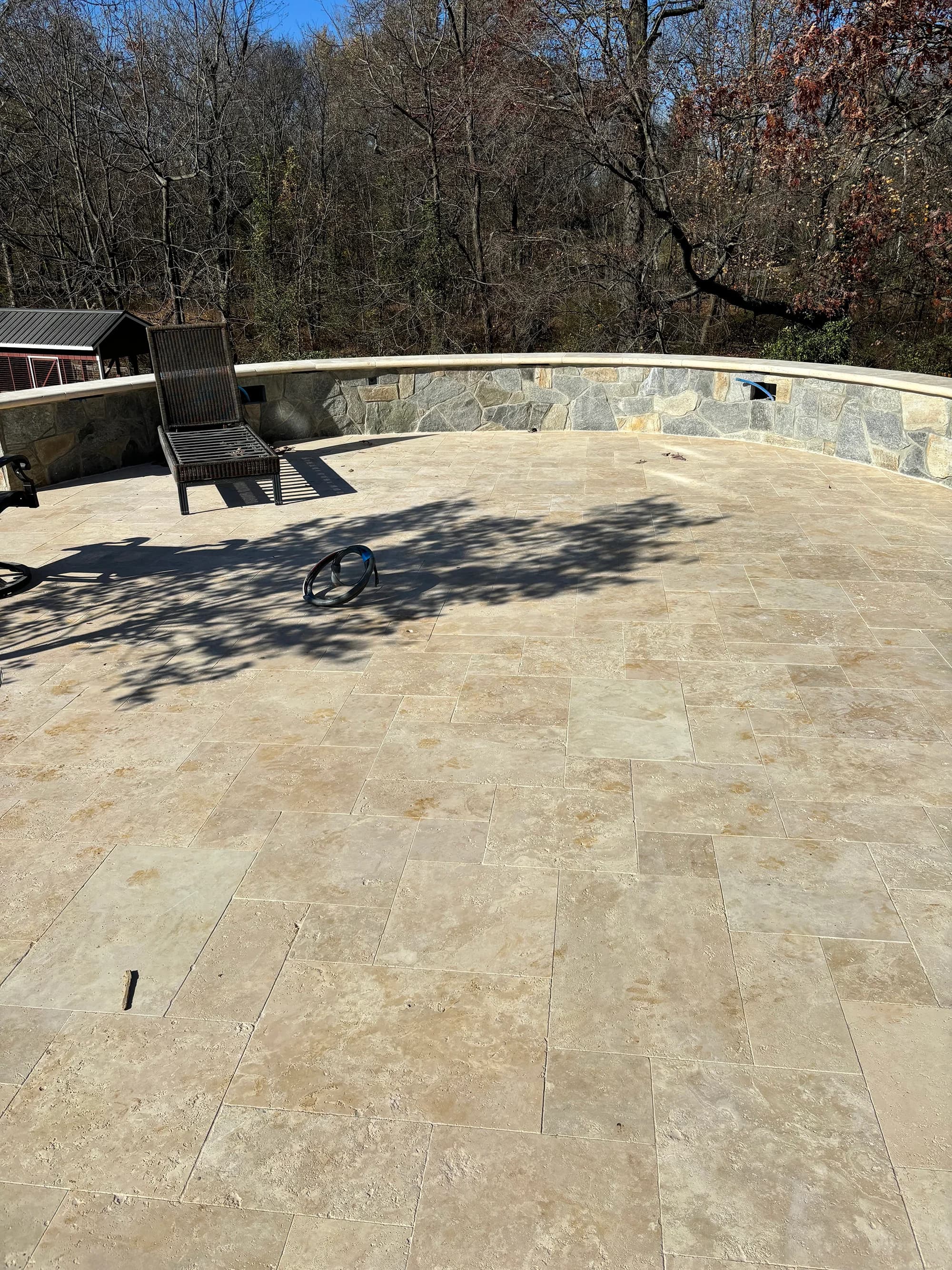 Ivory Travertine Tumbled French Pattern 3cm Paver - Select installed as a patio surface. The tumbled finish showcases a blend of soft beige tones with natural texture. Surrounded by a stone wall, the area features a lounge chair and overlooks a wooded ...