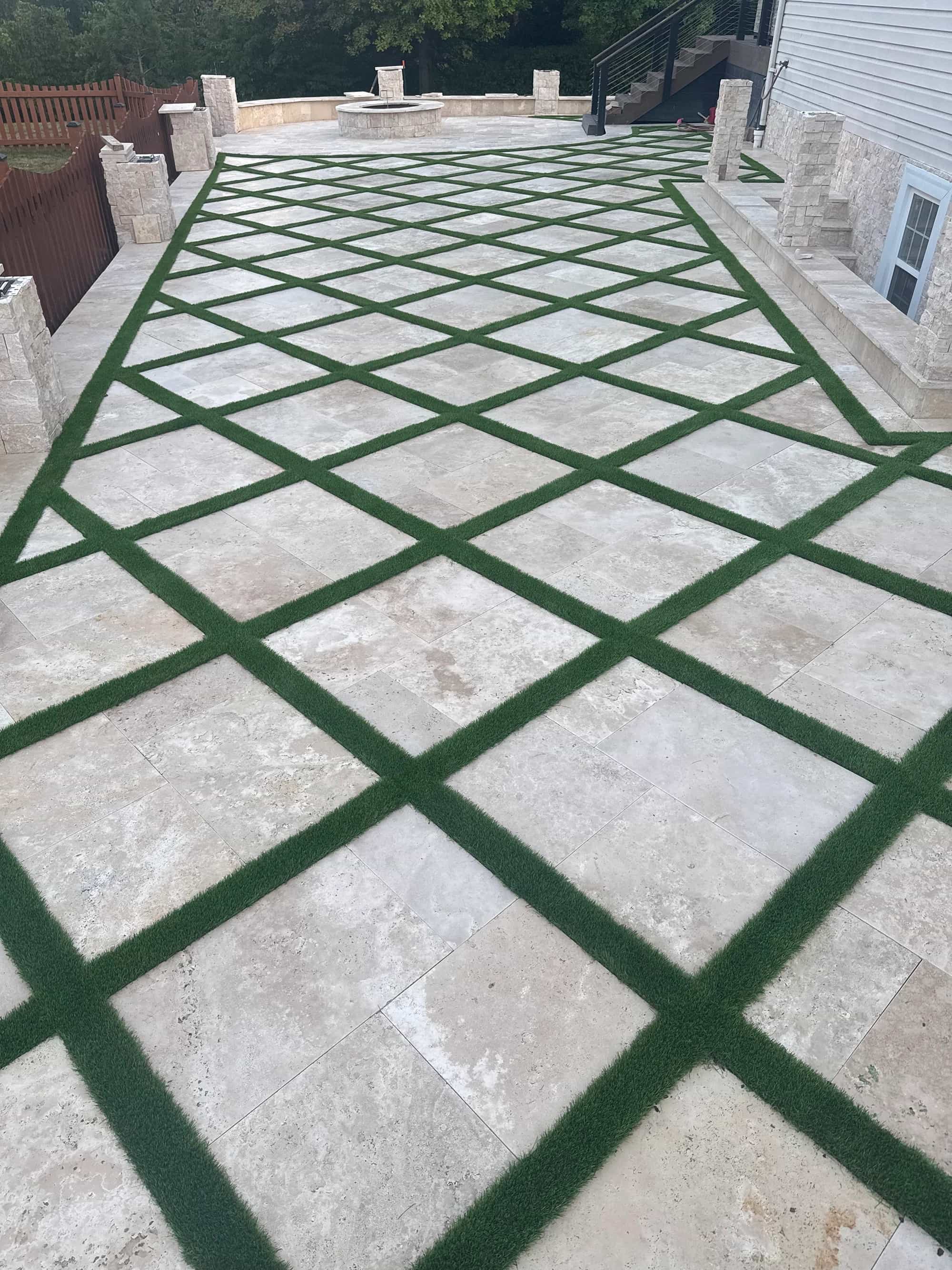 Ivory Travertine Tumbled French Pattern 3cm Paver installed in a patio with lush green grass accents. The natural stone's beige tones complement a modern outdoor space with stone walls, wooden fencing, and a central fire pit, creating a harmonious hard...