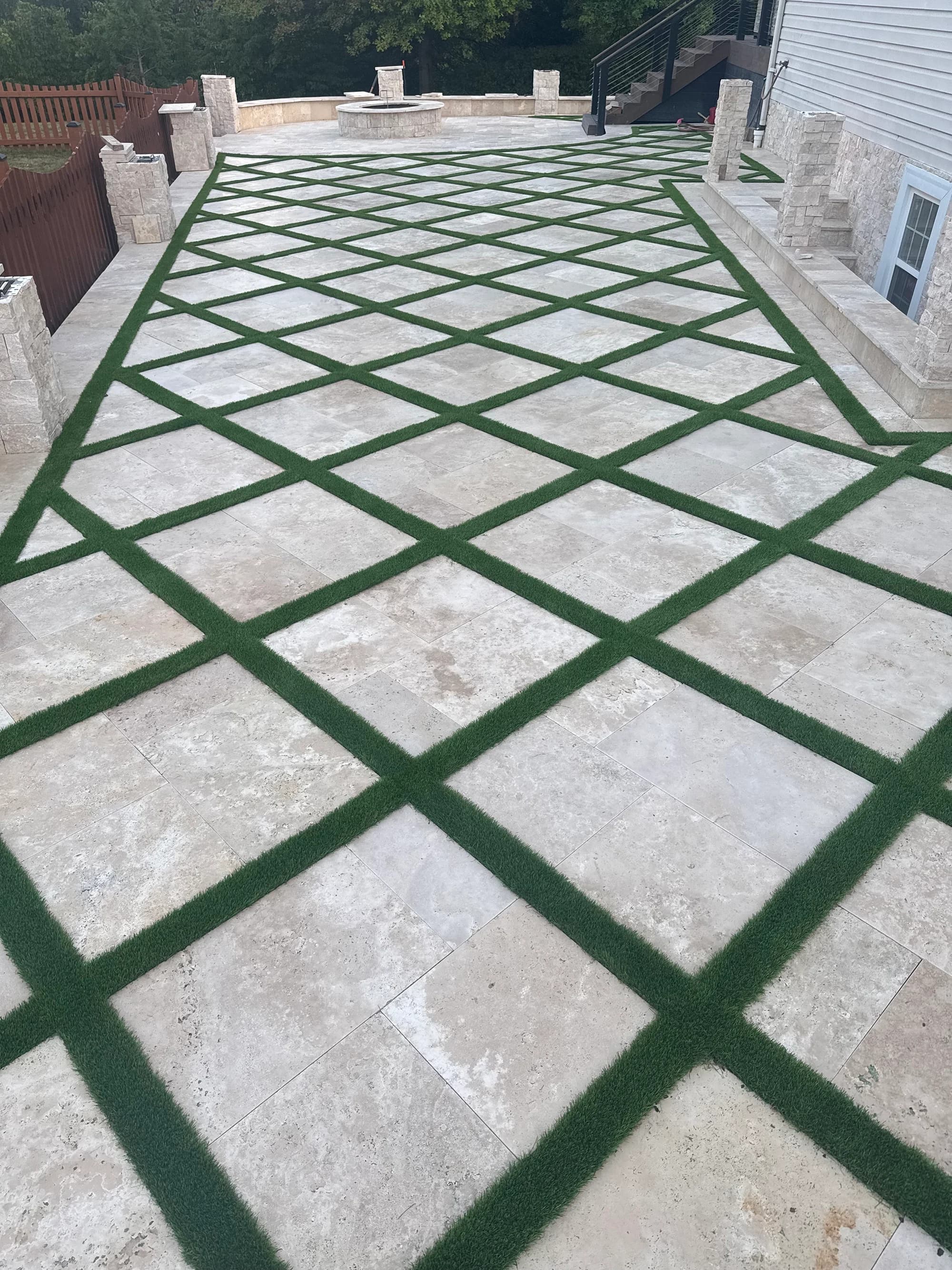 Ivory Travertine Tumbled French Pattern 3cm Paver installed in a patio with lush green grass accents. The natural stone's beige tones complement a modern outdoor space with stone walls, wooden fencing, and a central fire pit, creating a harmonious hard...