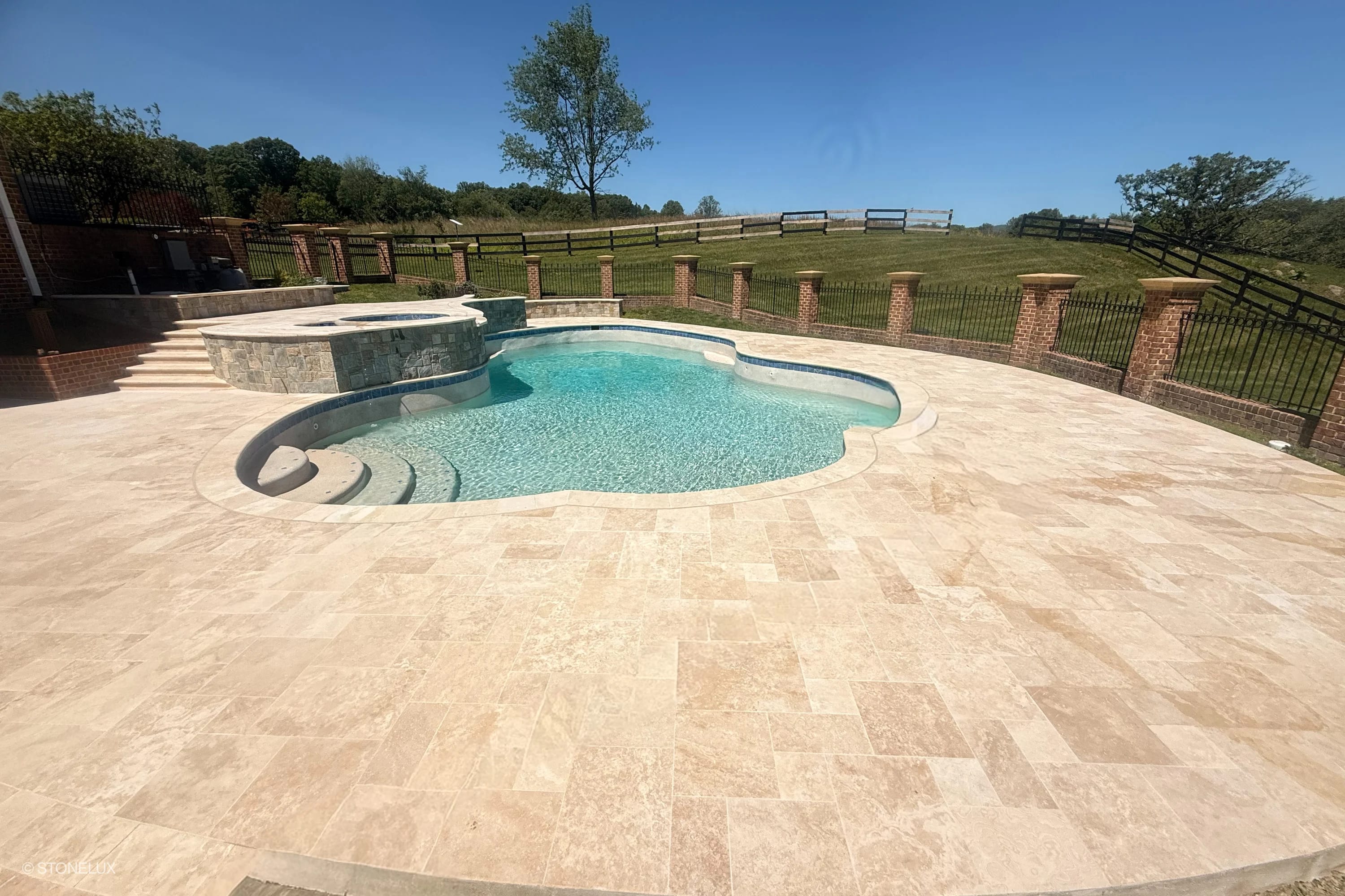 Ivory Travertine Tumbled French Pattern 3cm Pavers from STONELUX featured in a luxurious pool deck installation. The creamy, tumbled travertine complements the sparkling blue pool and rustic brick fencing, creating an elegant outdoor oasis.