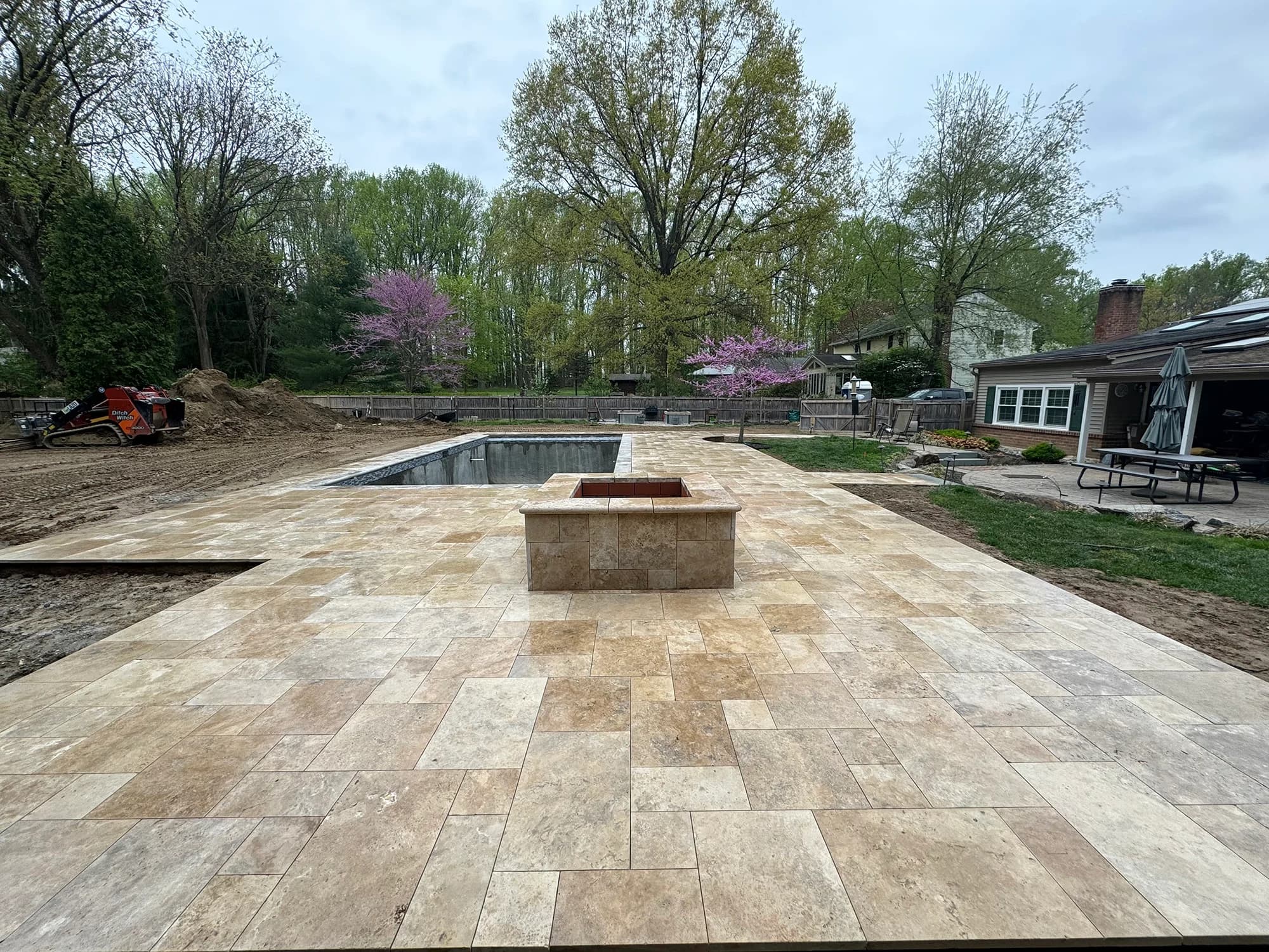 Outdoor installation showcasing Philly Travertine Tumbled French Pattern 3cm Paver by Stonelux. The natural stone patio features warm beige tones and a rustic finish, surrounding a pool construction site. Lush greenery and modern architecture complemen...